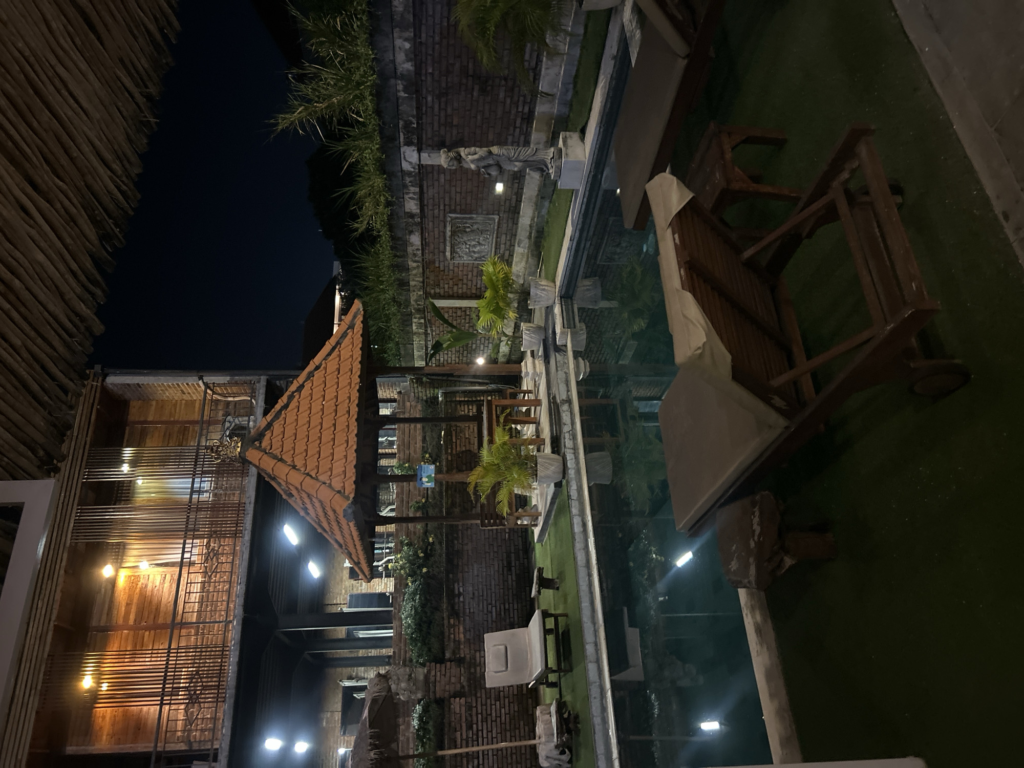 The image depicts an outdoor swimming pool area at night, likely in a resort or hotel, with a view of a multi-story wooden and brick building. The building features balconies with metal railings and illuminated windows. Below the building, a small open-air pavilion with a terracotta tiled roof is visible, furnished with a wooden table and chairs, surrounded by potted plants. Adjacent to the pavilion, a single beige lounge chair sits on artificial turf. A brick wall runs behind the pool area, adorned with a relief carving and a statue of a woman. Two wooden lounge chairs are positioned by the pool's edge. The pool itself is illuminated by underwater lights and the reflections of overhead lights. The overall scene is tranquil and suggestive of a tropical night setting. This scene is located in North Kuta, Indonesia.