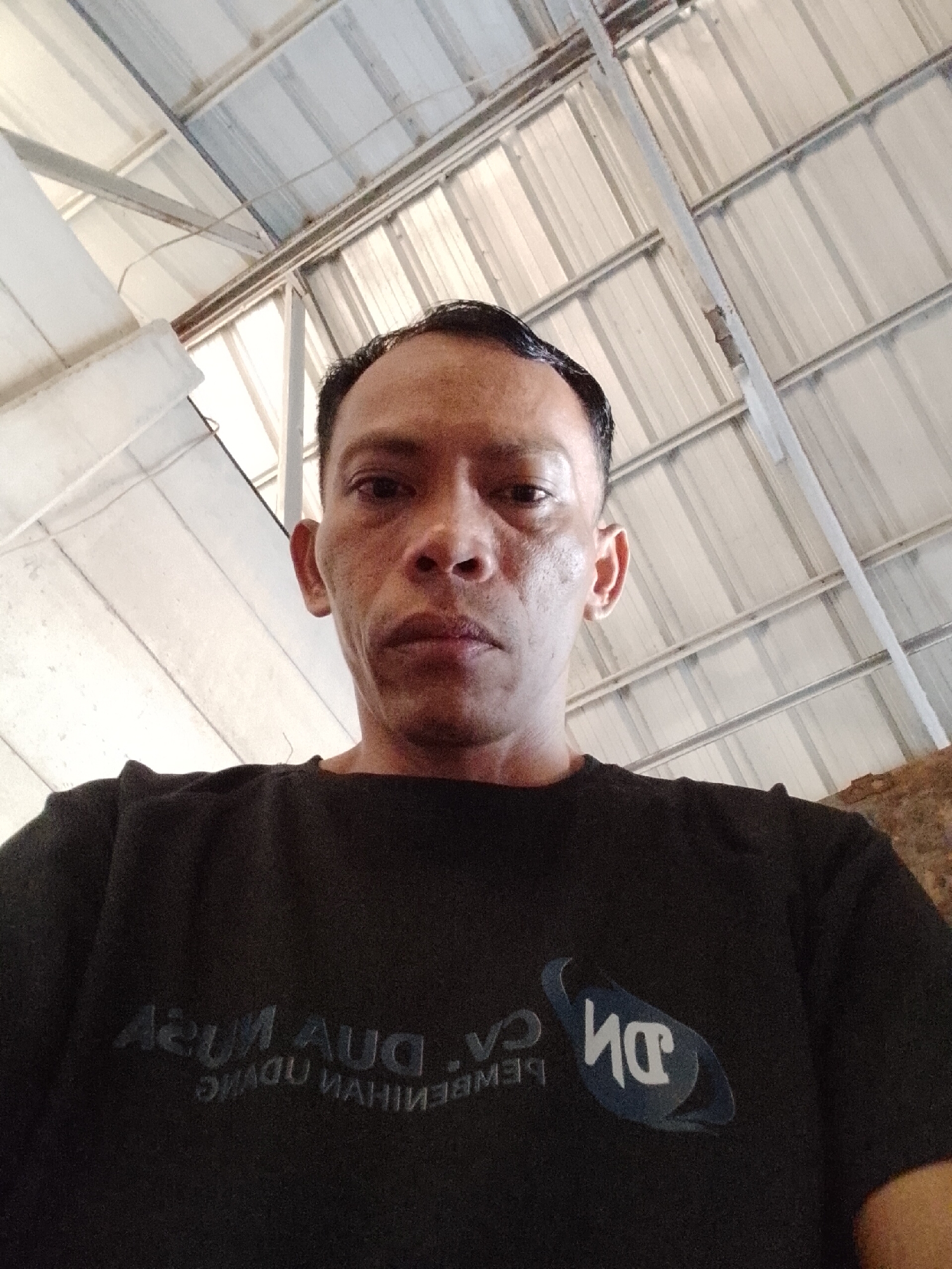 The image shows a close-up, low-angle selfie of a man in Bukit Ketok, Indonesia. The man is wearing a black t-shirt with a dark blue logo on the left side of his chest. The background is dominated by the corrugated metal roof of a structure, supported by white metal beams. The lighting suggests an indoor or covered outdoor environment. The man's face is the primary subject, with his expression neutral. The text on his shirt, which appears to be in a foreign language and is partially obscured, is also visible.