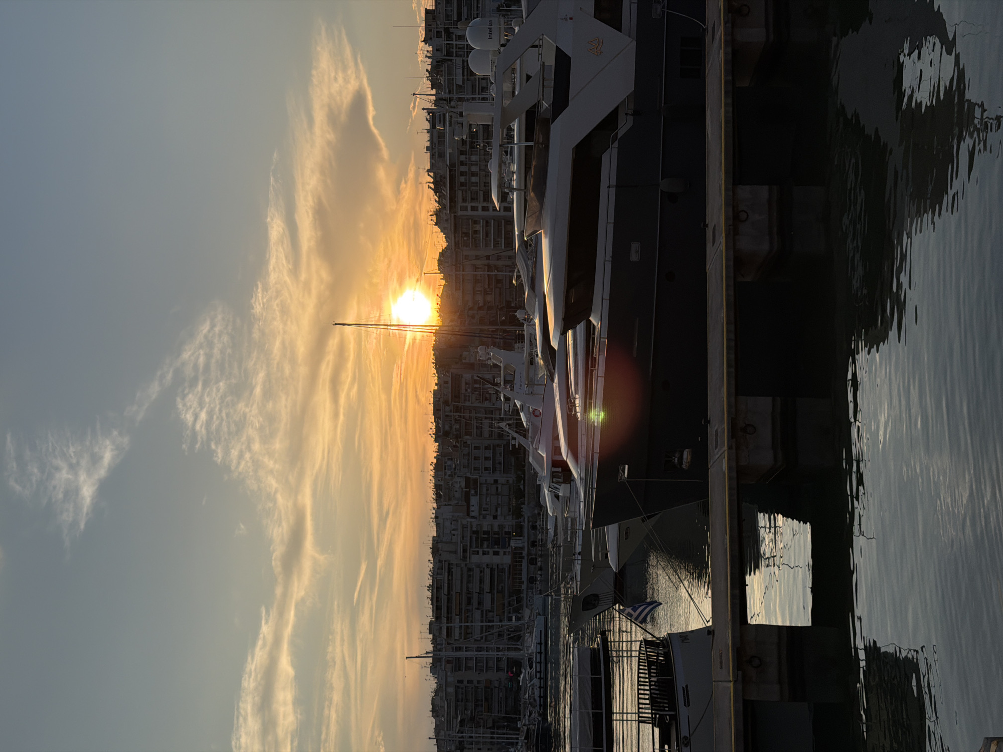 The image depicts a sunset over the harbor in Piraeus, Greece, with yachts docked along the pier. The sun is positioned in the center of the frame, partially obscured by buildings in the background and long wispy clouds. A mix of modern buildings line the shore. A dark-hulled yacht dominates the foreground on the right, and smaller boats are visible behind it. The water in the harbor reflects the light and colors of the sunset and boats. A Greek flag is visible on one of the smaller boats.
