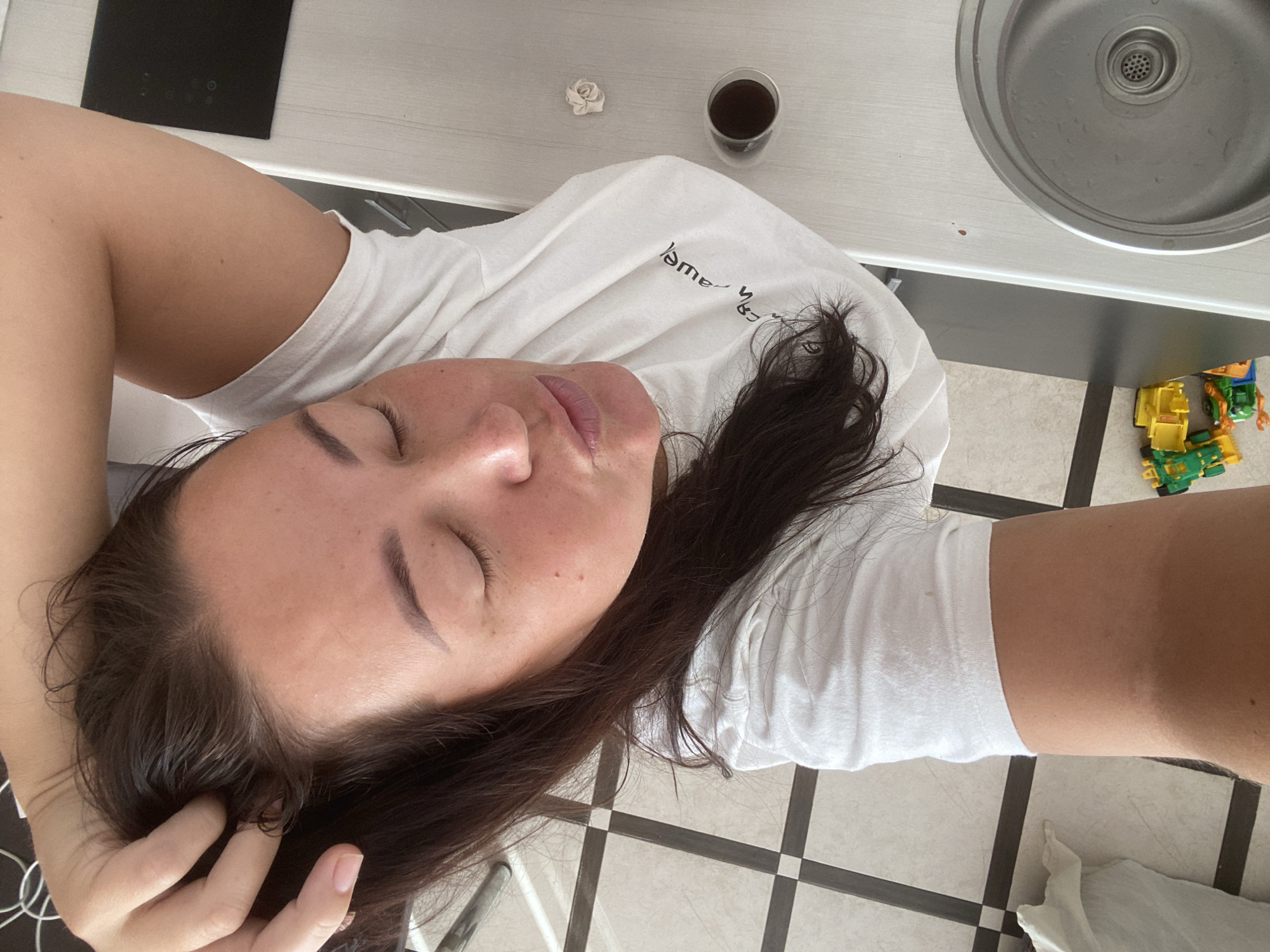 A woman with dark hair, eyes closed, and her lips pursed is lying down, her head propped on her arm. She is wearing a white t-shirt. The background shows a tiled floor and a metallic countertop. On the countertop, there is a glass of dark liquid and a small decorative item. The scene appears to be indoors, possibly a kitchen, with toys scattered on the floor. The image does not provide any information that would allow for an identification of the location within the city of Sochi, Russia.