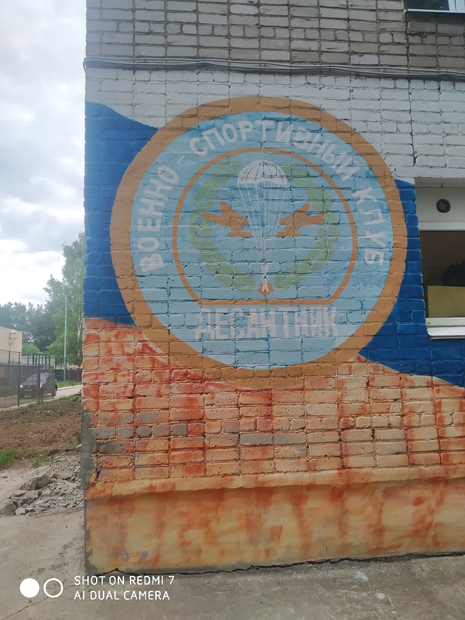The image shows a painted brick wall on the side of a building in Chaikovsky, Russia. The painting is a circular emblem, featuring the words "ВОЕННО-СПОРТИВНЫЙ КЛУБ" (Military Sports Club) and "ДЕСАНТНИК" (Paratrooper) in Cyrillic. Inside the circle, there is a drawing of a parachute flanked by two airplanes, with laurel wreaths at the bottom. The brickwork above the emblem is painted white, while the area around it is painted blue. The lower portion of the wall is painted in shades of orange and red, blending into the gray concrete ground. A window is visible on the right side of the wall. In the background, there are trees, a fence, and a glimpse of a building. A car is also visible near the fence. The lower left corner of the image contains the text "SHOT ON REDMI 7 AI DUAL CAMERA".