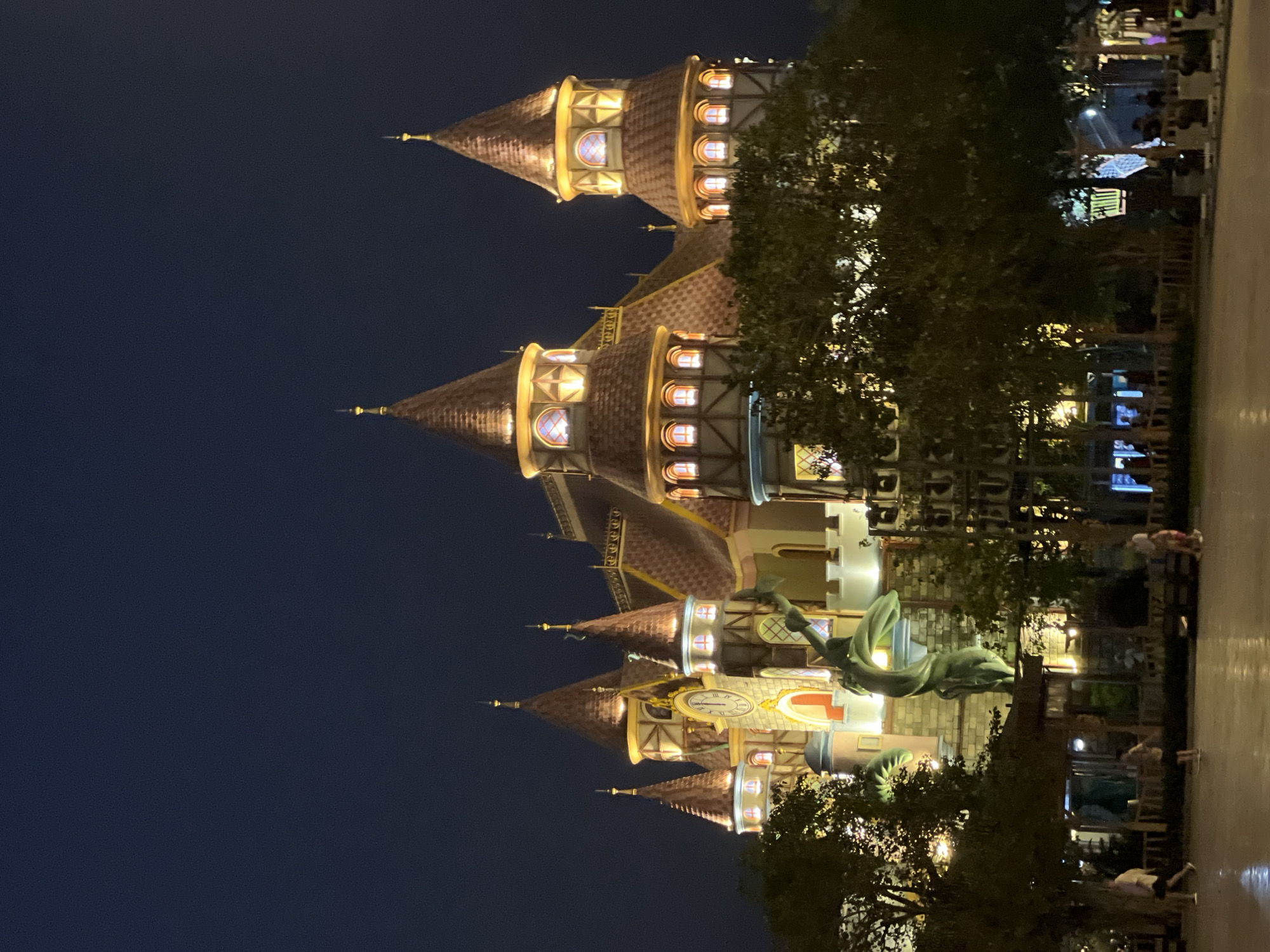 At night, a brightly lit castle-like structure stands as the focal point, possibly a tourist attraction in Nha Trang, Vietnam. The building has multiple towers with red-tiled roofs, many windows, and decorative detailing. A green dragon statue is visible near the base of the structure. People are walking on a walkway in the foreground, though their details are indistinct. Trees obscure parts of the building and the walkway. Streetlights and other light sources contribute to the overall illumination of the scene. The sky is dark and clear.