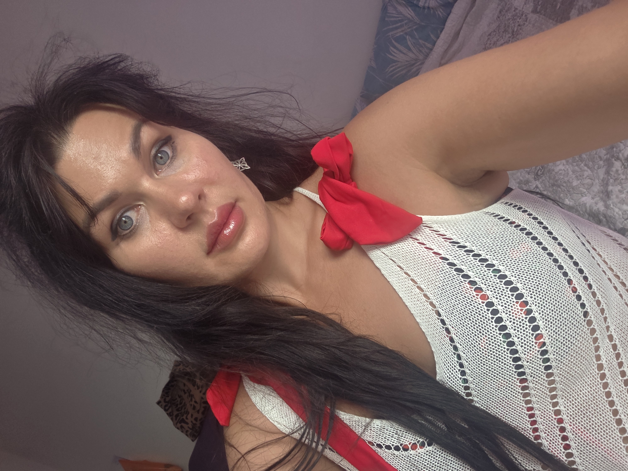 A selfie is taken from the chest up of a woman with long dark hair and light blue eyes. She is wearing a white, knitted top with red bows on the shoulders. She has on lip gloss and a silver earring in the shape of a star. She is leaning her head slightly to her left. The background is a wall with a patterned headboard and bedding. The scene is indoors and appears to be a bedroom. There is no indication of the city of Sochi, Russia.