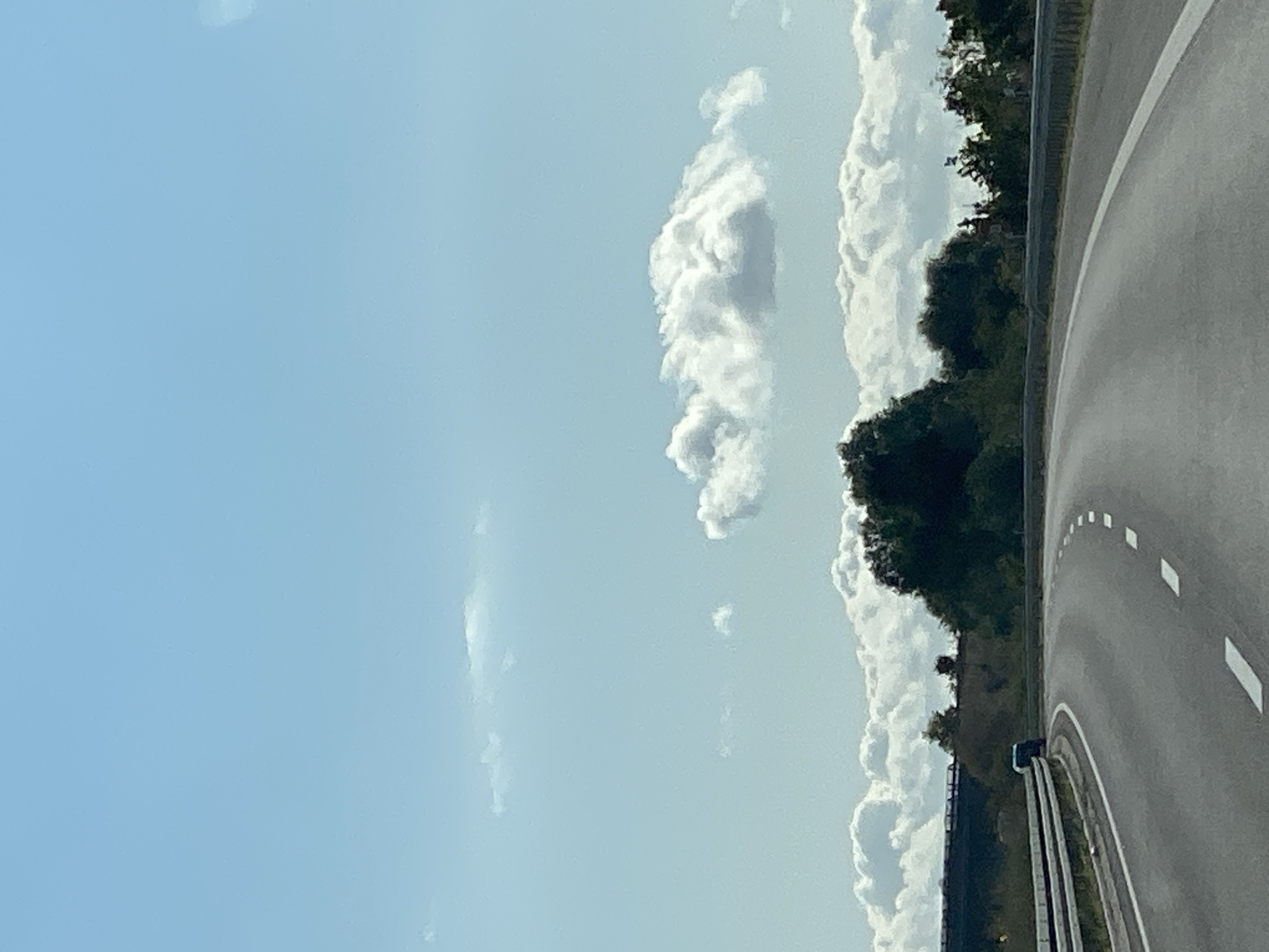An aerial view from a vehicle shows a highway curving to the right. A blue and white van is visible on the highway in the distance. To the right of the highway is a guardrail and a densely wooded area with various trees. In the background, a bridge is visible, and further back, a hazy skyline with a few buildings can be seen. The sky is a bright blue with scattered white clouds. The image does not provide any specific information about the city of Heringen/Helme, Germany.