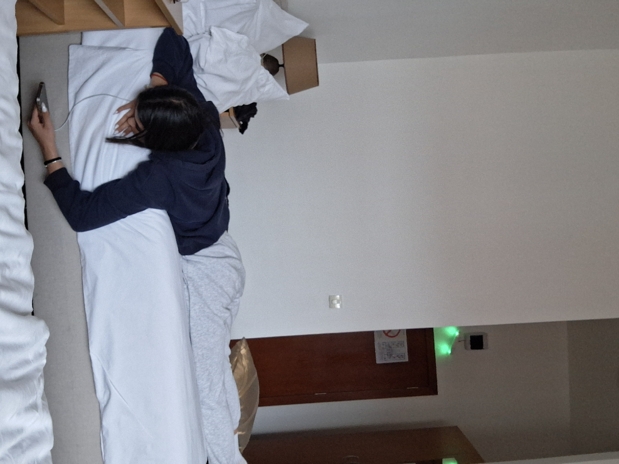 The scene depicts an interior room identified as being within the city of Vozdovac Urban Municipality, Serbia, likely a hotel or dormitory. A single individual is positioned prone on a bed covered with white linens and a white duvet, their head turned away from the viewer or face down. The individual wears a dark blue long-sleeved top and light gray trousers. Their right hand, extended downward, holds a dark-colored mobile phone connected to a white cable, possibly a charging cable or wired earphones. The left hand rests near the individual's face, also appearing to interact with a white cable.
To the left of the bed, a wooden bedside table is visible, topped with a table lamp featuring a brown conical shade. Behind the lamp, white pillows are stacked. To the right, a dark brown wooden door with a metallic handle is set into a light-colored wall. A white rectangular sign, bearing a red "No Smoking" pictogram and additional indecipherable text, is affixed to the door. Above this door, a series of small green lights, possibly decorative or indicator lights, are visible. Further right, a section of a dark wooden wardrobe or another door is partially visible. The walls are uniformly light-colored. A golden-colored pillow is partially visible on the bed towards the foot.