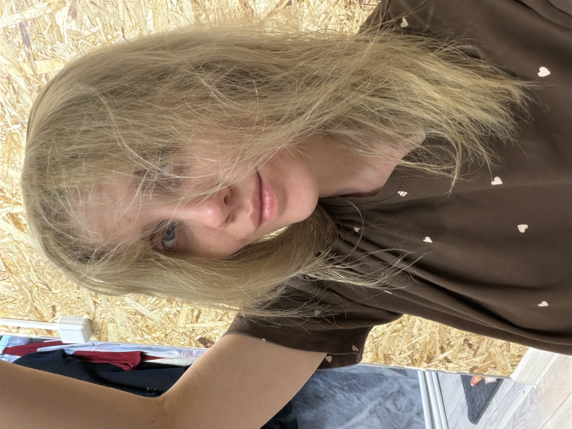 A selfie of a blond woman is presented, partially obscured by her own hair. She has fair skin and is wearing a brown top with a subtle pattern of small white hearts. The background appears to be a wall or paneling constructed from wood chips. In the lower part of the frame, there are hints of clothing items, including what appears to be a red garment. The angle of the photo suggests it was taken from above, with the camera tilted. Specific location in the city of Chaikovsky, Russia cannot be determined from the image.