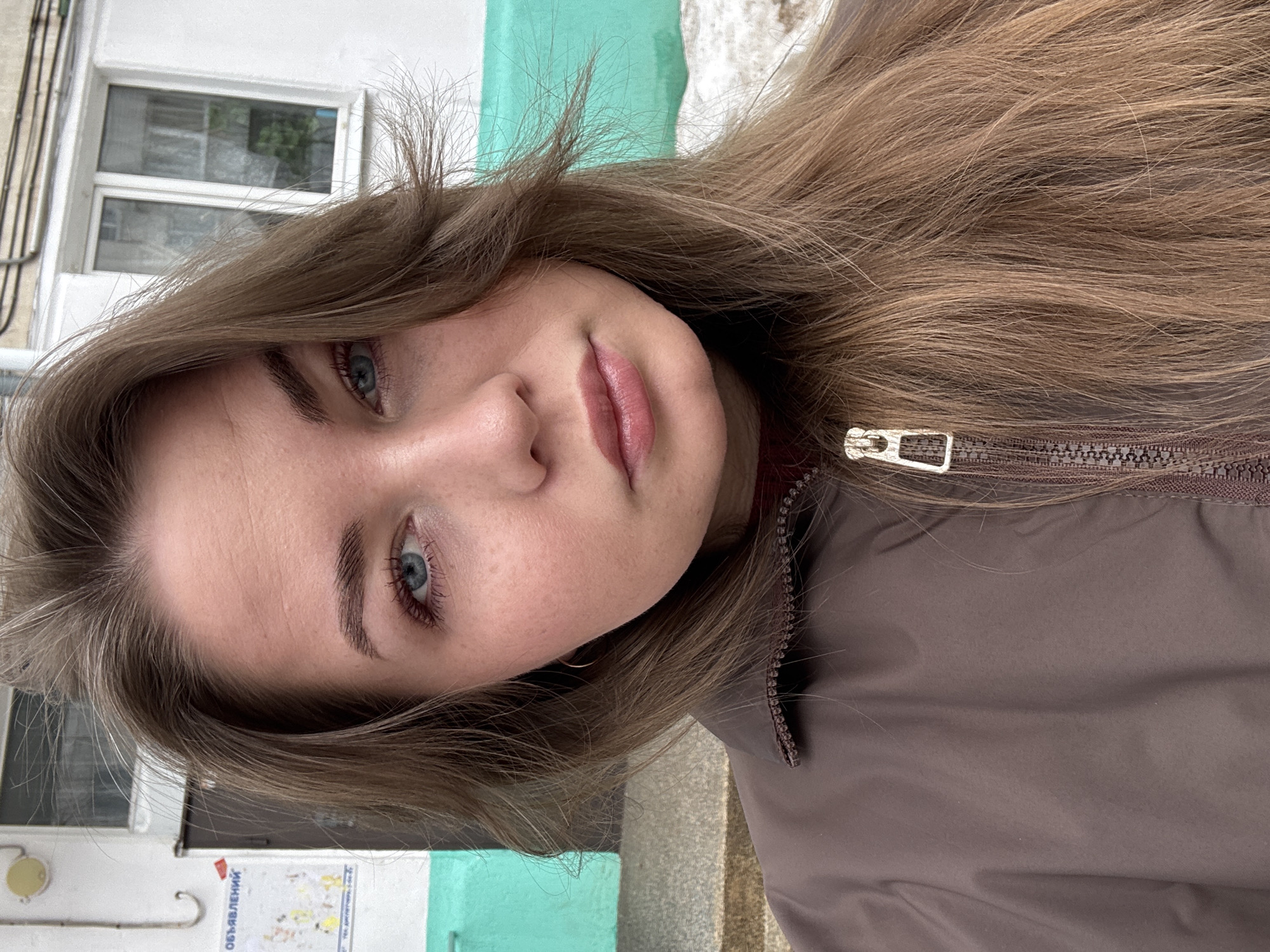 The image is a close-up selfie of a female individual, approximately 20-30 years old, with long, light brown hair and blue eyes, captured outdoors in what appears to be a residential area of Chaikovsky, Russia. She wears a brown zippered jacket, with a dark red collar visible underneath, and has a neutral expression with slightly parted lips. In the background, a two-tone building features a white upper section and a bright teal/turquoise lower section. A dark brown entrance door is visible on the left, above which a white bulletin board with the Cyrillic word "ОБЪЯВЛЕНИЯ" (Announcements) is affixed. To the right of the door, a white-framed window with two panes is set into the white wall. Pipes or conduits are visible running horizontally across the white wall section. A circular light fixture or sensor is mounted on the wall above the doorframe. The ground includes concrete steps on the left and patches of dirty snow on the far right, indicating a cold season such as late winter or early spring.