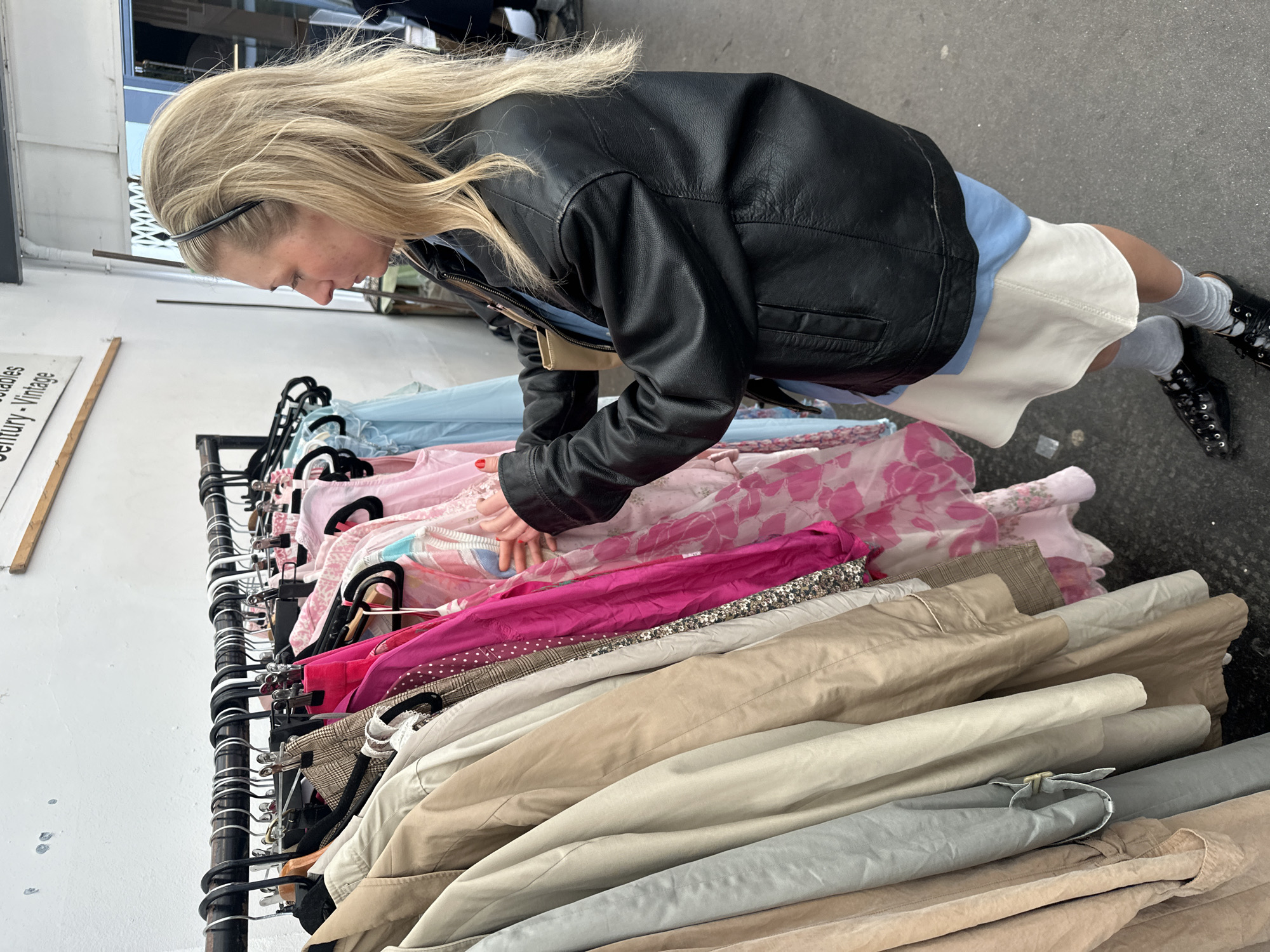 The image depicts a blonde woman in a black leather jacket browsing a rack of clothes at what appears to be a vintage or second-hand clothing sale. The woman is leaning over the rack, examining the garments. The rack is densely packed with items, ranging from pastel-colored tops to beige jackets. A sign is partly visible on the left side of the image and contains the words "Ladies Century Vintage". The setting implies an outdoor or semi-outdoor market or fair, suggestive of a street in a city. The location cannot be specifically identified as London from the given image.
