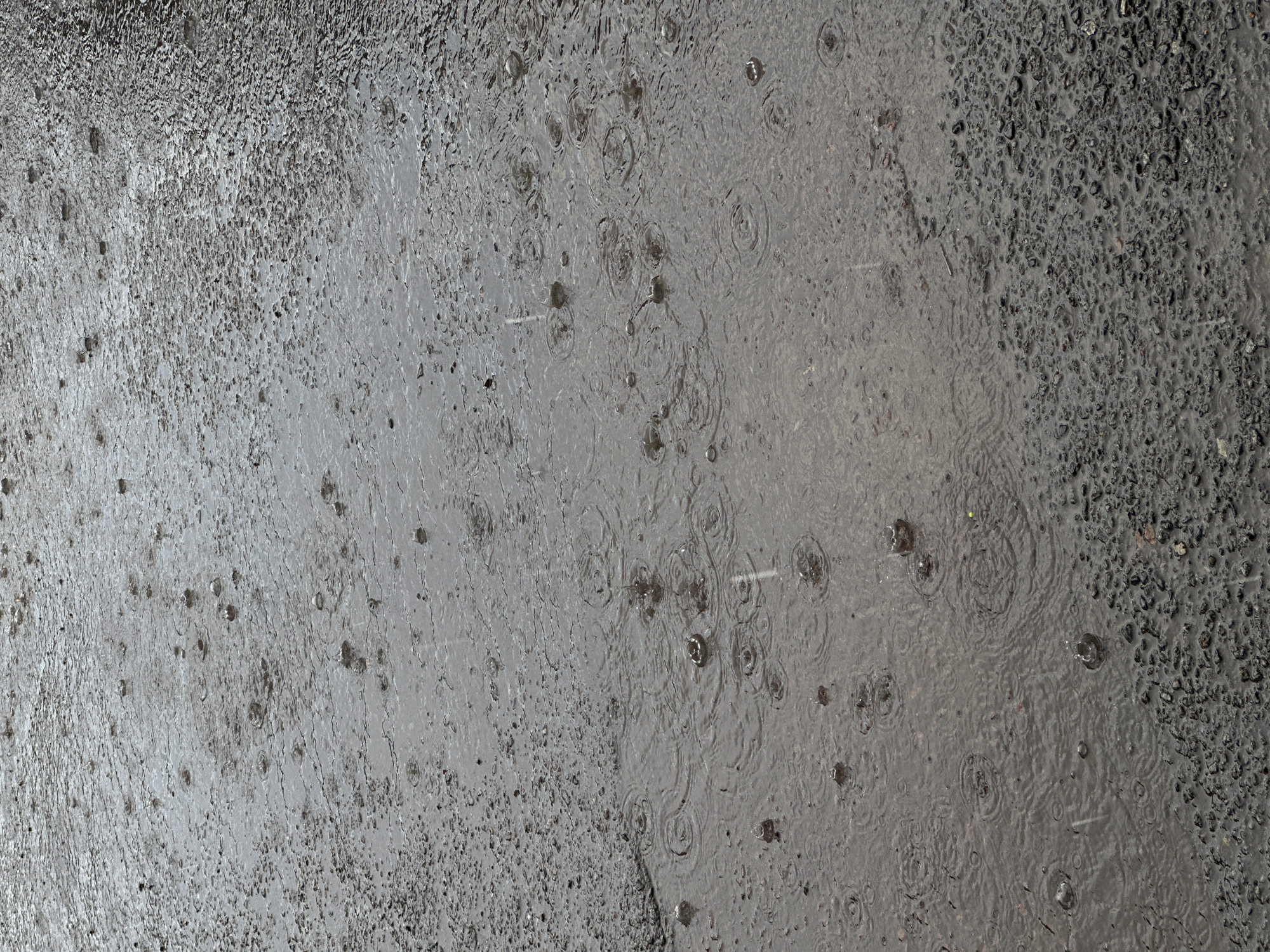 The image depicts an outdoor scene of rainfall on a rough asphalt surface. Small ripples and splashes are visible on the wet surface, indicating active precipitation. The asphalt appears to be a mixture of dark aggregate and finer material, with some areas showing a smoother, waterlogged texture and others a more granular appearance. The lighting is diffused, suggesting an overcast or rainy sky. There are no people or discernible structures in the frame. The setting is likely an urban environment, possibly a street or walkway, in Kokshetau, Kazakhstan, given the provided context.