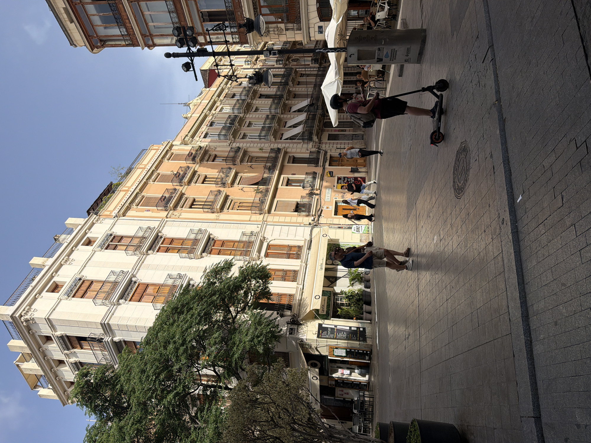 The image depicts a city square with traditional European architecture and pedestrians. In the foreground, a person is riding an electric scooter. To the left, two people are walking and conversing. Further back, several other individuals are visible, some walking and others seated at outdoor cafe tables. The buildings surrounding the square feature multiple stories with balconies and ornate detailing, suggesting a historical urban setting. The ground is paved with cobblestones. A large tree stands to the left, providing shade. The sky is clear and blue.