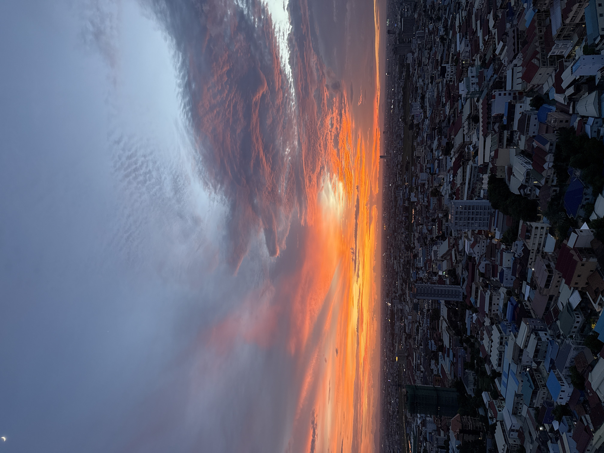 This high-angle, full shot captures a vibrant sunset over the urban landscape of Khan Mean Chey, Cambodia. The sky is a spectacle of color, transitioning from a deep blue at the top to brilliant oranges and reds near the horizon, with wispy clouds catching the last rays of sunlight. A thin crescent moon is visible in the upper left quadrant of the sky. Below the dramatic sky, a sprawling city unfolds, characterized by densely packed residential buildings with a variety of roof colors, including red, blue, and white. Several taller buildings, including a partially constructed skyscraper covered in green netting, punctuate the cityscape. The lights of the city are beginning to twinkle as dusk settles, indicating the scene is captured during twilight. No people or specific actions are discernible from this vantage point. The environment is clearly an urban setting with a dense population evident in the multitude of buildings.