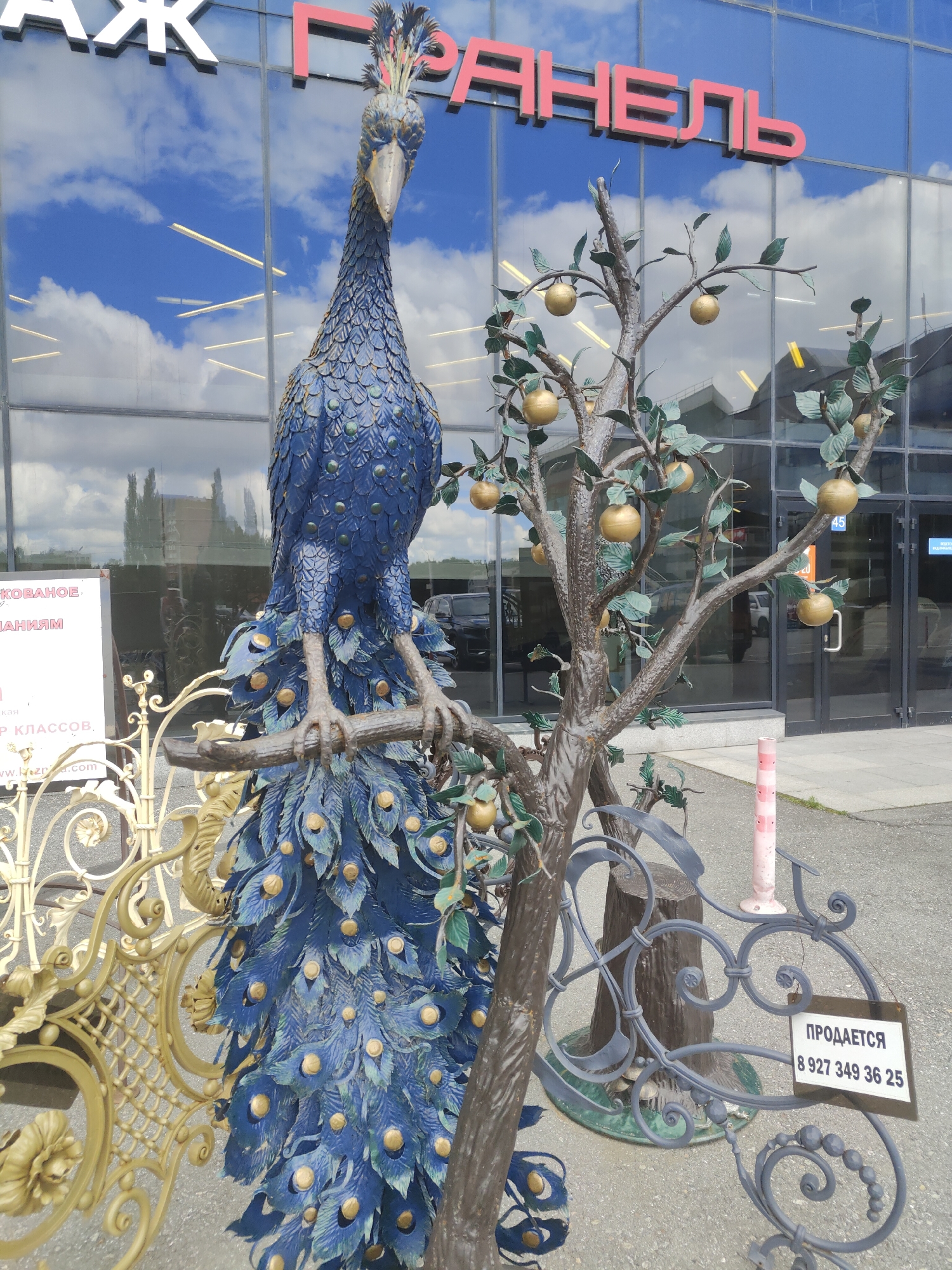 The image shows a statue of a blue peacock standing on a branch, with a metal tree bearing golden apples beside it. The statue is positioned in front of a building with a glass facade, reflecting the sky and clouds. There are also various metal decorative elements, a white sign with Russian text, and a pink and white striped pole. In the background, cars are visible parked near the building. The ground is paved with asphalt. The scene appears to be outdoors, in a public area. The text on the sign "ПРОДАЕТСЯ" translates to "FOR SALE" and a phone number is listed below it.