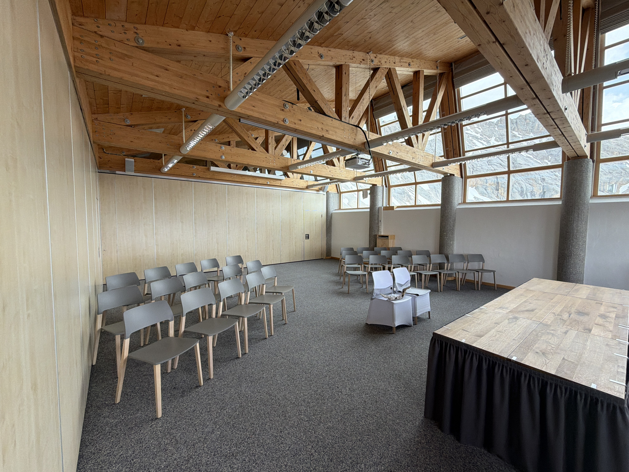 A photograph depicts an interior space that appears to be a conference or event venue, likely located in a mountainous region due to the landscape visible through the windows.
The room features a high, exposed wooden beam ceiling with multiple industrial-style lighting fixtures. Large windows span the far wall, offering a view of a mountain range possibly in or near Garmisch-Partenkirchen, Germany. The natural light illuminates the interior.
The foreground contains a stage with a wooden top and a black draped skirt. Rows of gray chairs are arranged for audience seating, predominantly on the left side of the frame. Towards the middle, there are two small tables covered in white cloths.
The color palette is neutral, with the warm tones of the wood contrasting with the cool grays of the chairs and carpet. The walls are a light beige, contributing to the bright and airy atmosphere of the space. There are no people visible in the shot.