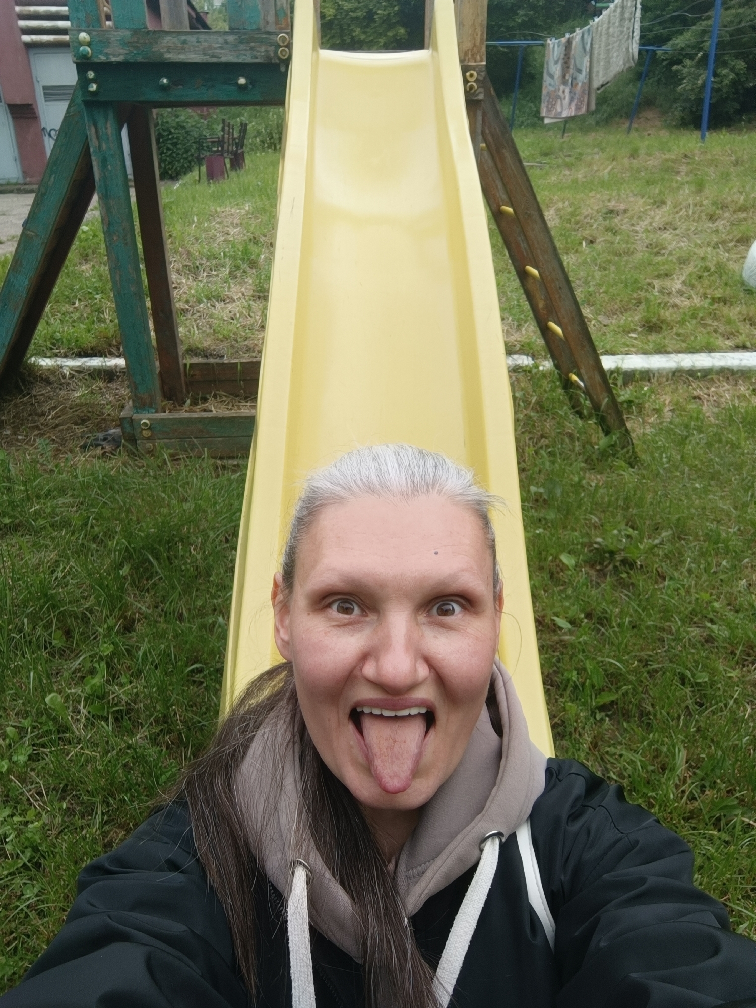 A woman is taking a selfie on a yellow slide, sticking out her tongue and widening her eyes. She has grey hair and is wearing a black jacket with a grey hoodie underneath. The slide is part of a playground structure, with green-painted wooden supports on the left and brown wooden supports on the right. Green grass is visible in the background. Clothes are hanging on a clothesline in the background. This image was taken outdoors. There is no indication that the image was taken in the city of Lermontov, Russia.