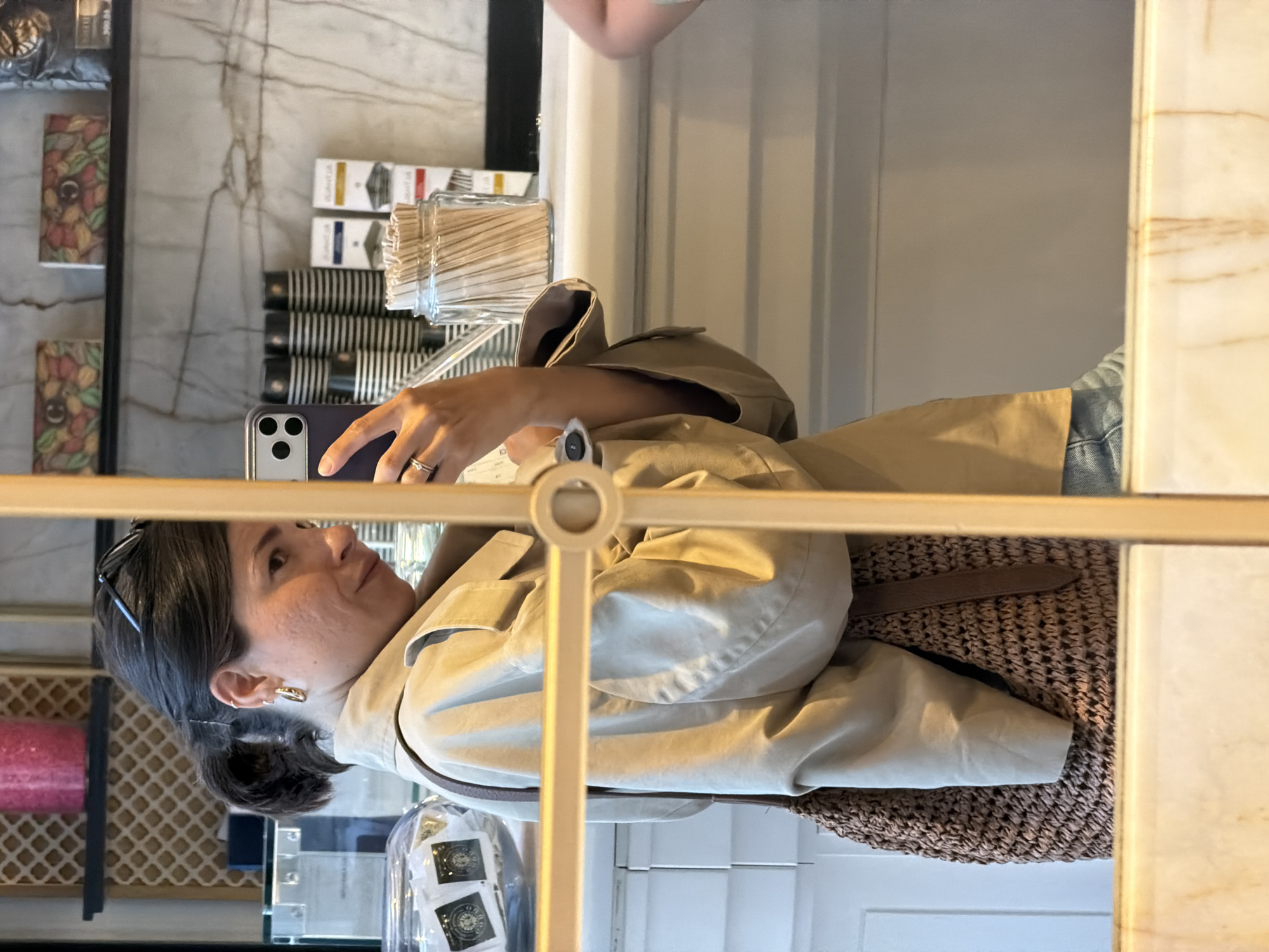 A woman is taking a picture with a smartphone in what appears to be a café or store. The woman is wearing a light brown trench coat, has dark hair pulled back in a ponytail, and is wearing gold hoop earrings. She is holding the smartphone in her left hand, with a ring visible on her finger. The background includes shelves stocked with products like tea, coffee cups, and a jar of wooden stirrers. There is a glass or plexiglass barrier in the foreground, partially obscuring the view. The location appears to be in Budva, Montenegro.