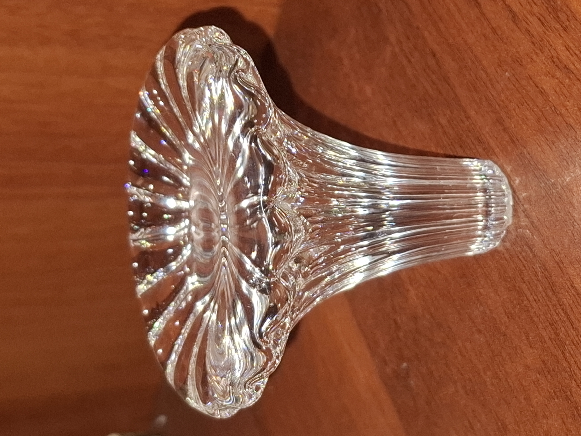 A clear glass object with a fluted design, resembling a mushroom cap, rests on a wooden surface. The object is illuminated, causing light to refract and create rainbow-like glints on its surface. The background is a warm, reddish-brown color, consistent with wood. The scene appears to be indoors, possibly in a home or display area. There is no indication of the city of Almaty, Kazakhstan.