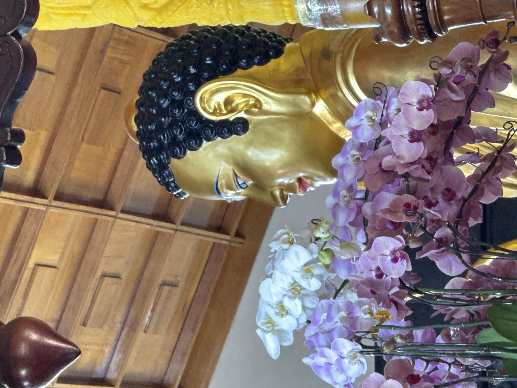 The image depicts a close-up, side profile of a golden Buddha statue adorned with a wreath of purple and white orchids. The Buddha's head is covered in dark, curly hair, and its face is serene with closed eyes. The statue is positioned indoors, with a wooden ceiling and a decorative wooden element visible in the upper portion of the frame. To the right of the Buddha, a yellow, textured pillar or decorative element is partially visible. The scene likely captures a devotional offering or decorative arrangement within a Buddhist temple or a similar spiritual site. The presence of orchids suggests a tropical or subtropical environment, consistent with Da Lat, Vietnam, which is known for its floral cultivation and temperate climate.