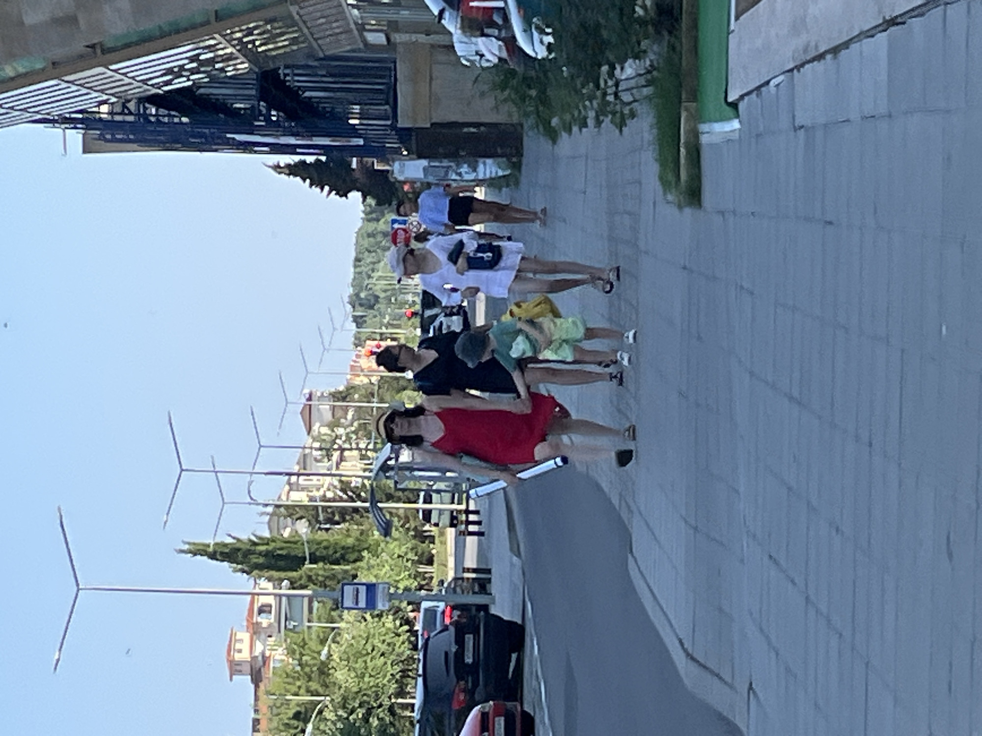 A group of people are walking along a sidewalk in Nesebar, Bulgaria. In the foreground, a woman in a red dress is holding a rolled-up poster and walking with a young boy in a green shirt and shorts. Behind them, a woman in a black dress is walking, and further back, a woman in a white dress and a woman in a blue shirt are also walking. The sidewalk is paved with gray tiles. To the left of the sidewalk is a road with parked cars. To the right of the sidewalk is a modern building with a glass facade and some greenery. The background shows trees and buildings with red roofs. Several streetlights are visible along the road.