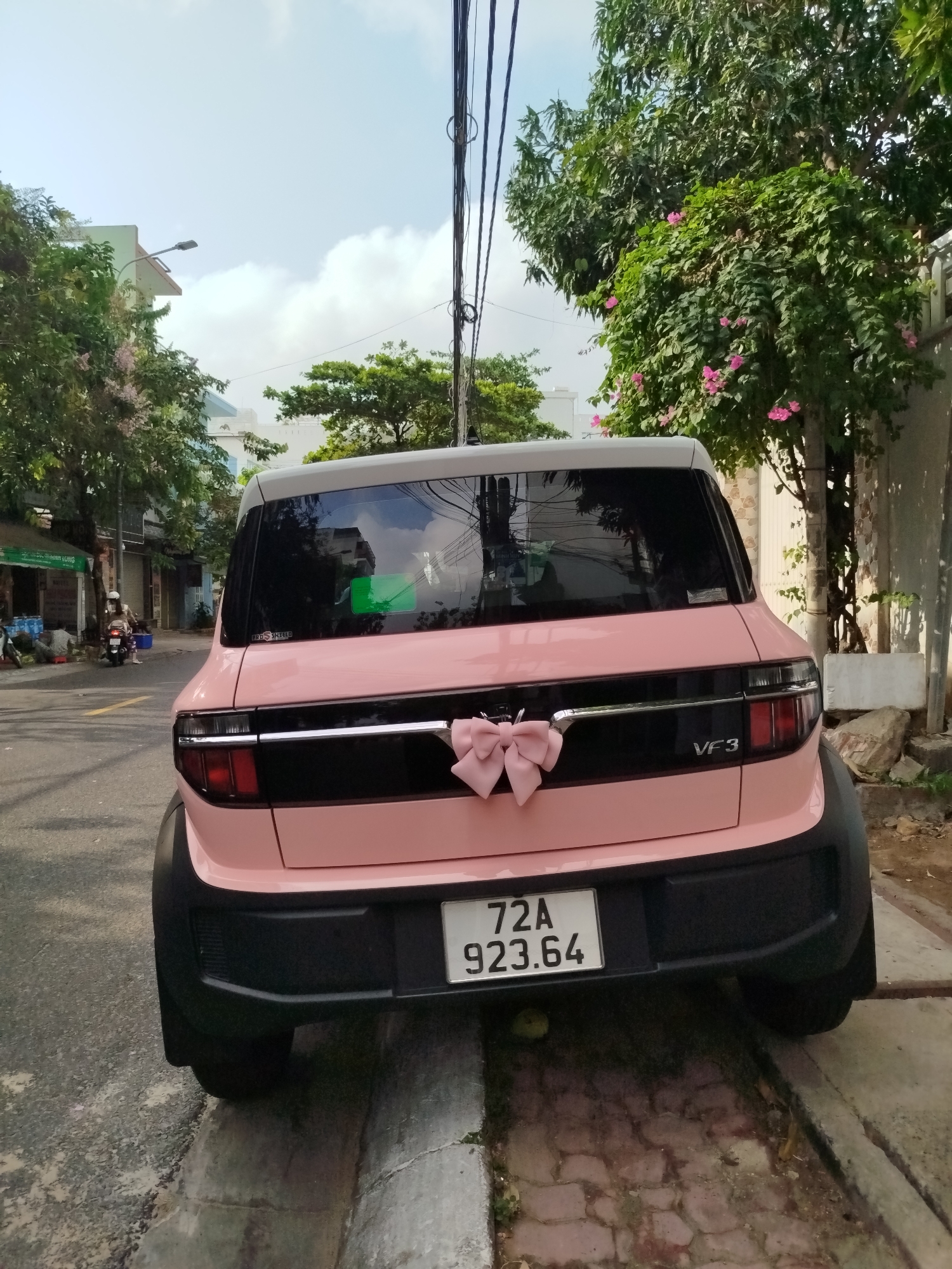 A pink VinFast VF3 electric car is parked on a street in Ho Chi Minh City, Vietnam. The rear of the car faces the camera. A light pink bow is attached to the chrome trim on the tailgate. The license plate number 72A 923.64 is visible. The car is parked near the curb. Trees and buildings are visible in the background, along with utility lines. A person on a motorbike can be seen in the distance on the left side of the frame.