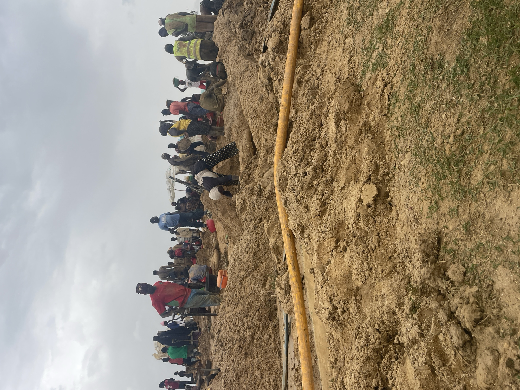 The image shows a large group of people congregated on a dirt embankment or hillside, likely in Maiyanga, Nigeria. The ground is bare earth, sparsely covered with grass in some areas. A yellow pipe runs vertically along the side of the embankment. The sky is overcast. Individuals are dressed in a variety of clothing, with some wearing work vests or carrying items on their heads. Some people are standing on top of each other.