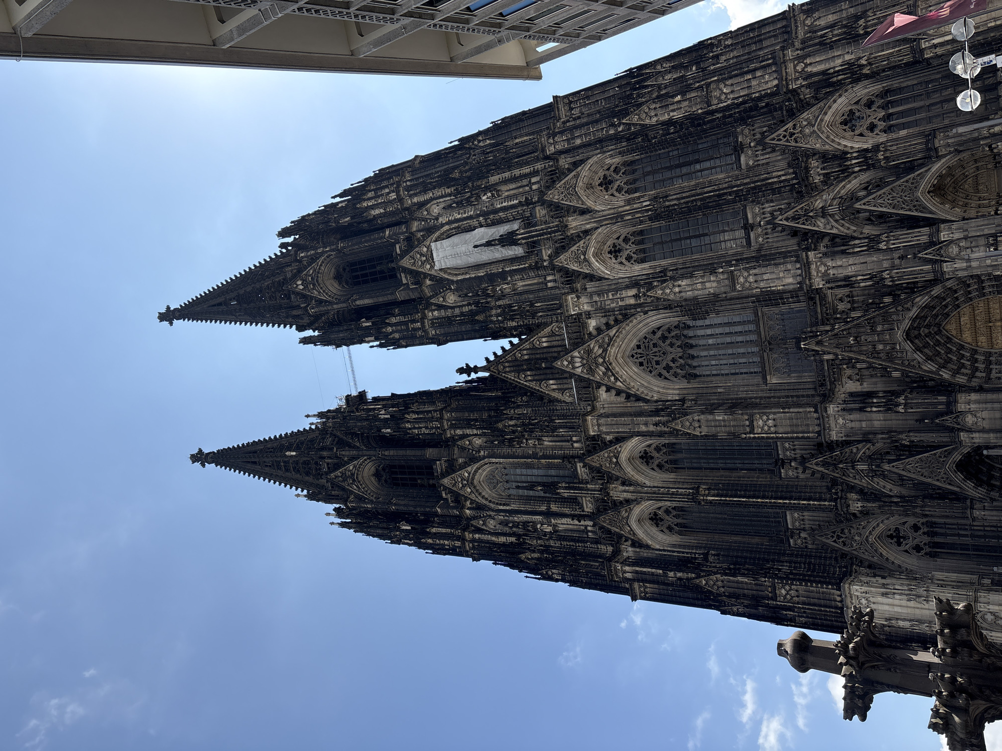 The image shows a low-angle view of the Cologne Cathedral, also known as Kölner Dom, a prominent landmark in Cologne, Germany. The cathedral's twin spires dominate the frame, reaching towards a clear blue sky with a few wispy clouds. The intricate Gothic architecture of the cathedral, characterized by its dark stone facade, pointed arches, and numerous spires and pinnacles, is clearly visible. Scaffolding and netting are present on one of the spires, indicating ongoing restoration or maintenance work. To the right of the cathedral, a modern building with a light-colored facade and vertical windows is partially visible, contrasting with the historical structure. In the lower left corner, a section of a gargoyle-adorned stone structure, likely part of the cathedral's exterior, is visible. The lighting suggests it is daytime, with sunlight casting shadows and highlighting the architectural details.