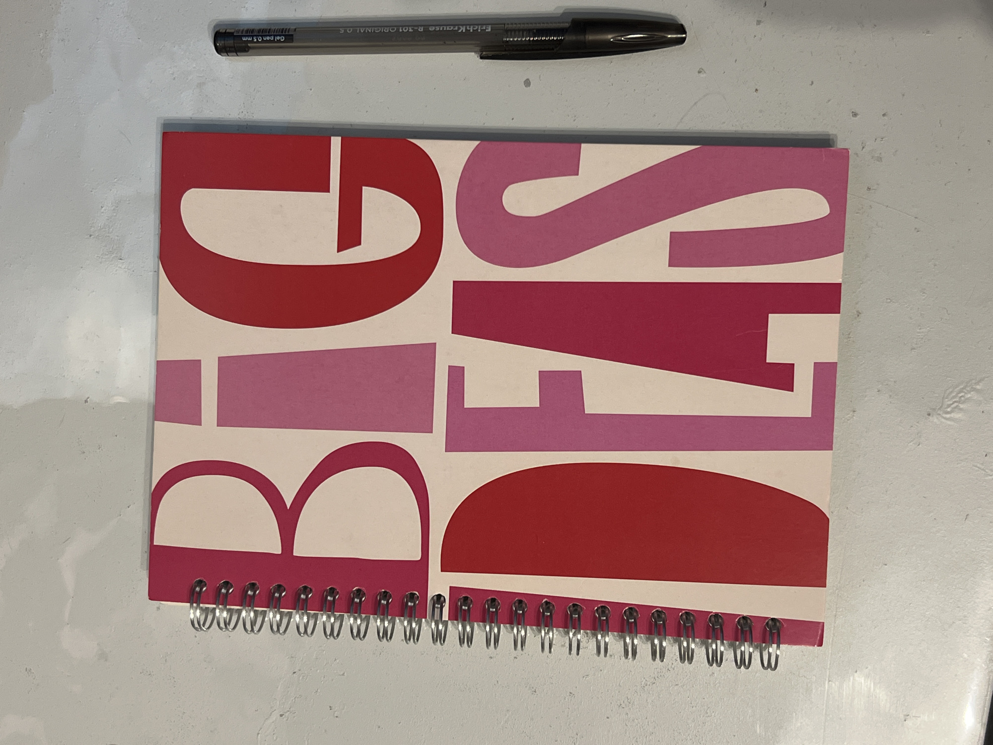 A flat lay image features a black ErichKrause R-301 ORIGINAL 0.5 gel pen positioned horizontally above a spiral-bound notebook. The pen's barrel clearly displays the text "Gel pen 0.5 mm".
The notebook cover is adorned with large, stylized block letters in shades of red and pink on an off-white background. The word "BIG" is fully visible on the left side of the notebook, primarily in a deep red. To its right, the word "IDEAS" is partially visible, with letters "I", "D", "E", "A", and parts of "S" appearing in a mix of lighter pink and red hues. The notebook is bound along its bottom edge with a silver-colored metal spiral.
The objects rest on a light grey, textured surface, possibly a concrete floor or a weathered table. The lighting is even and diffused, suggesting an indoor setting. No human activity is depicted; the image is a static arrangement of everyday objects. While the location context is Kazan, Russia, no specific visual cues within the image itself indicate this geographical location.