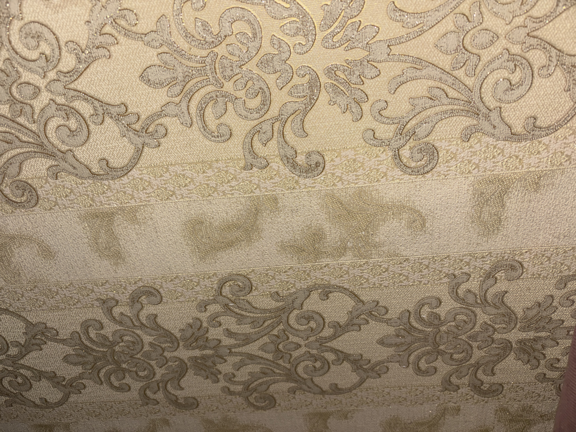 The image displays a close-up of textured wallpaper with a damask pattern. The wallpaper is primarily a pale gold or cream color, with raised swirling designs in a darker beige or taupe. There are vertical stripes of a lighter, almost glittery texture separating the main patterned sections. The pattern itself consists of ornate, baroque-style motifs with scrolling leaves and floral elements. The lighting in the image appears to be indoor, casting subtle shadows that highlight the texture of the wallpaper. The scene is static and focuses solely on the decorative wallpaper. There is no indication of any specific location within Sultan-Yangi-Yurt, Russia, as the image exclusively shows the texture and pattern of the wallpaper.