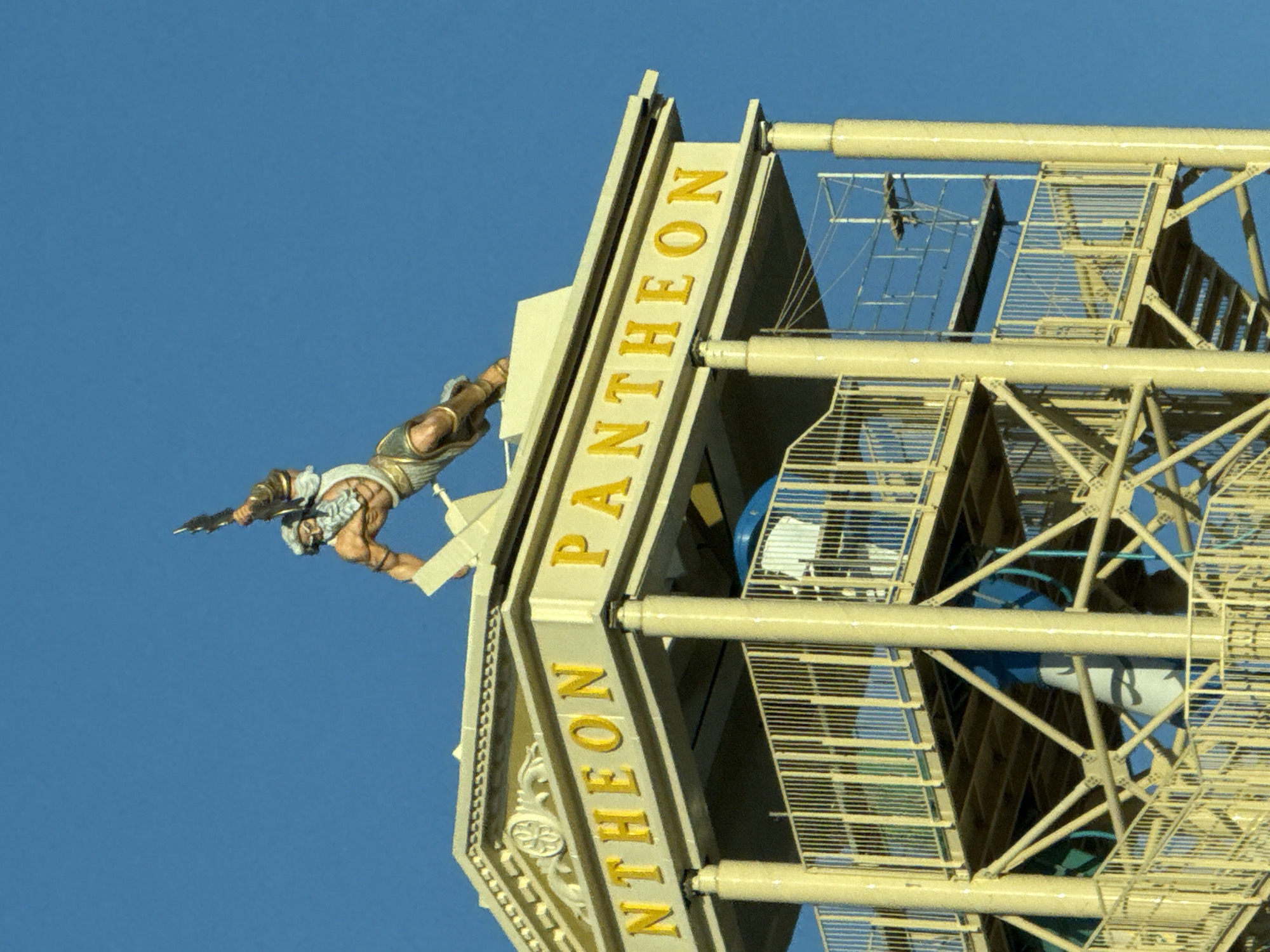 The image shows a close-up view of the upper section of a tower-like structure against a clear blue sky. At the very top of the structure is a statue of a muscular male figure, likely a god or hero from Greek mythology, poised as if in action, holding a weapon. The statue is painted in realistic tones of brown, white, and gold.
Below the statue is a pedimented roof-like structure with decorative carvings and the golden text "PANTHEON" prominently displayed. The architecture appears to be inspired by classical Greek design, with columns supporting the roof.
The rest of the image is dominated by the tower itself, which is constructed with a yellow metal framework. Multiple levels are visible, featuring walkways with yellow railings and what appear to be sections of a water slide with blue and white elements visible through the structure. Scaffolding is also present on one side of the tower. The overall impression is of an amusement park attraction or a themed building. The location is identified as Chersonisos Municipal Unit, Greece.