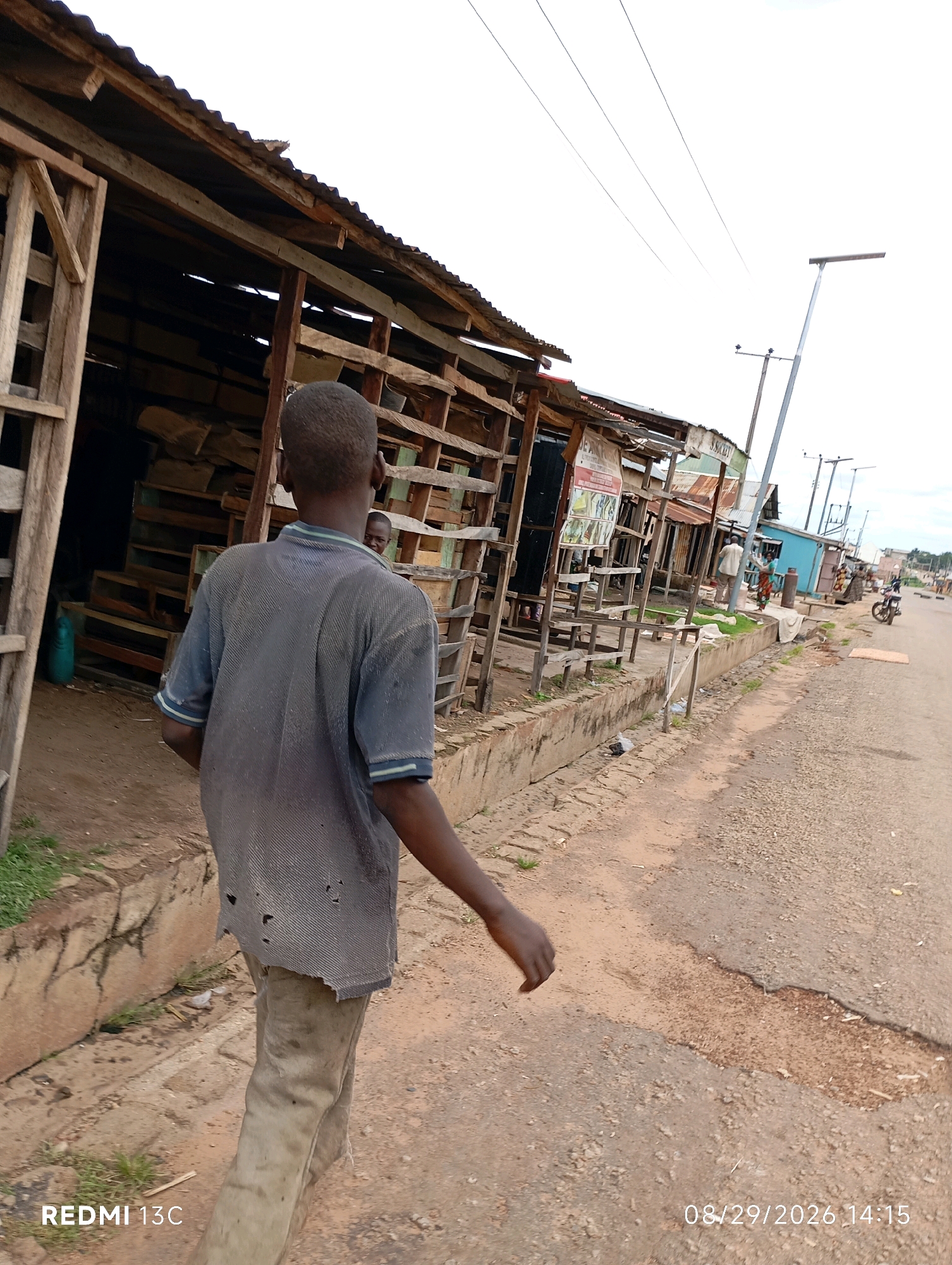 In Shendam, Nigeria, a young person walks away from the camera on a dirt road alongside a row of makeshift market stalls. The stalls, constructed from wood and corrugated metal, appear weathered and worn. The ground is a mix of dirt and gravel, with a concrete drainage ditch running alongside the road. The person is wearing a tattered grey shirt and light-colored pants. In the background, other people can be seen near the stalls, and a motorcycle is parked on the right side of the road. The sky is overcast.