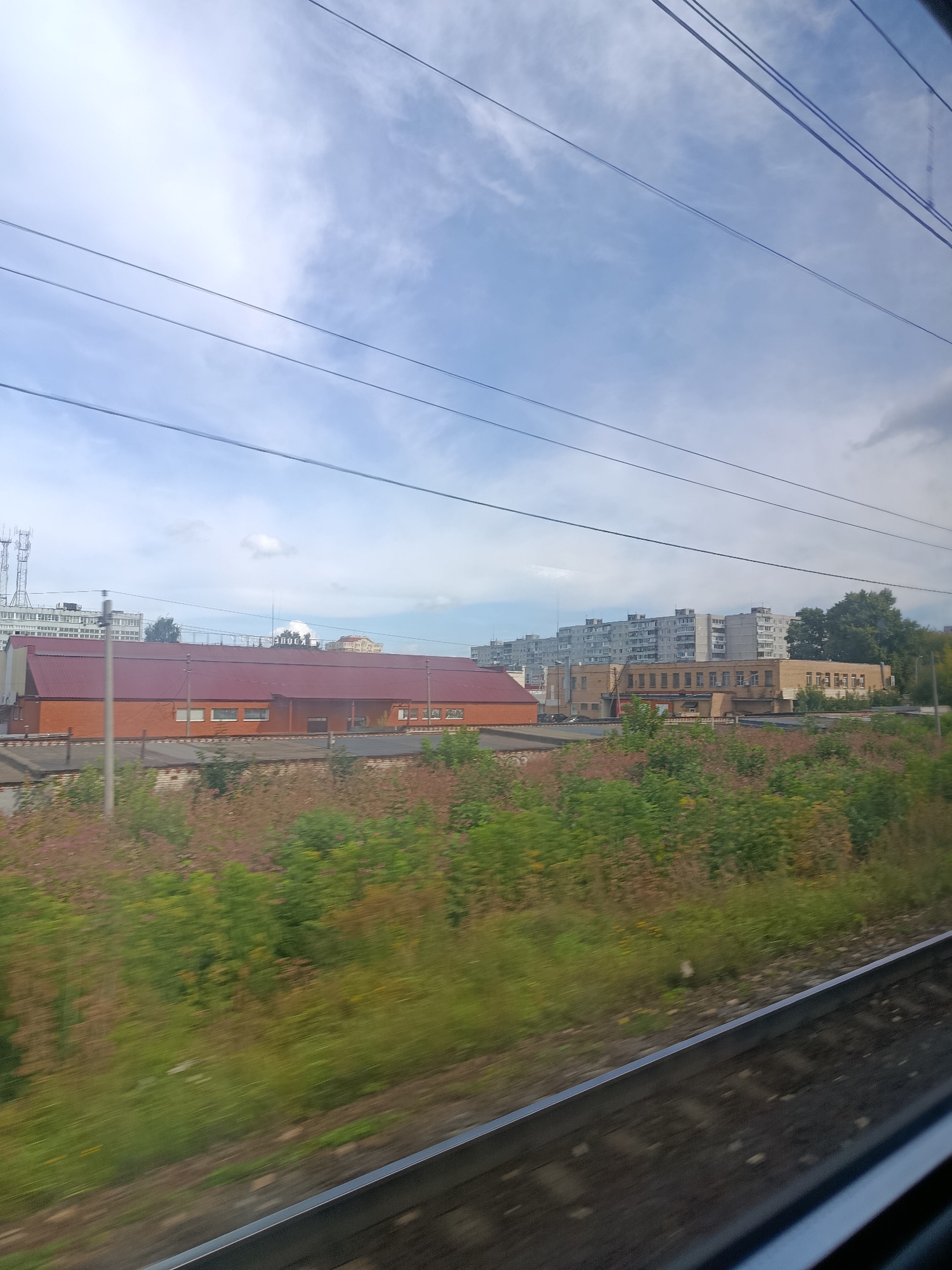 The image captures a view from a moving train, likely on the outskirts of Pavlovsky Posad, Russia, given the provided context. The foreground shows blurred train tracks and a grassy embankment with scattered pink and green vegetation. In the midground, a long, low building with a red-tiled roof and orange brick facade stretches horizontally. Adjacent to this structure, more industrial-looking buildings, also primarily brick, are visible. Further in the background, a cluster of tall, grey apartment buildings dominates the skyline, characteristic of Soviet-era urban planning. The sky above is partly cloudy with a mix of blue sky and greyish clouds, and several power lines crisscross the frame. The overall impression is that of an industrial or semi-urban landscape seen from a transportation corridor. There are no visible people or animals in the image.