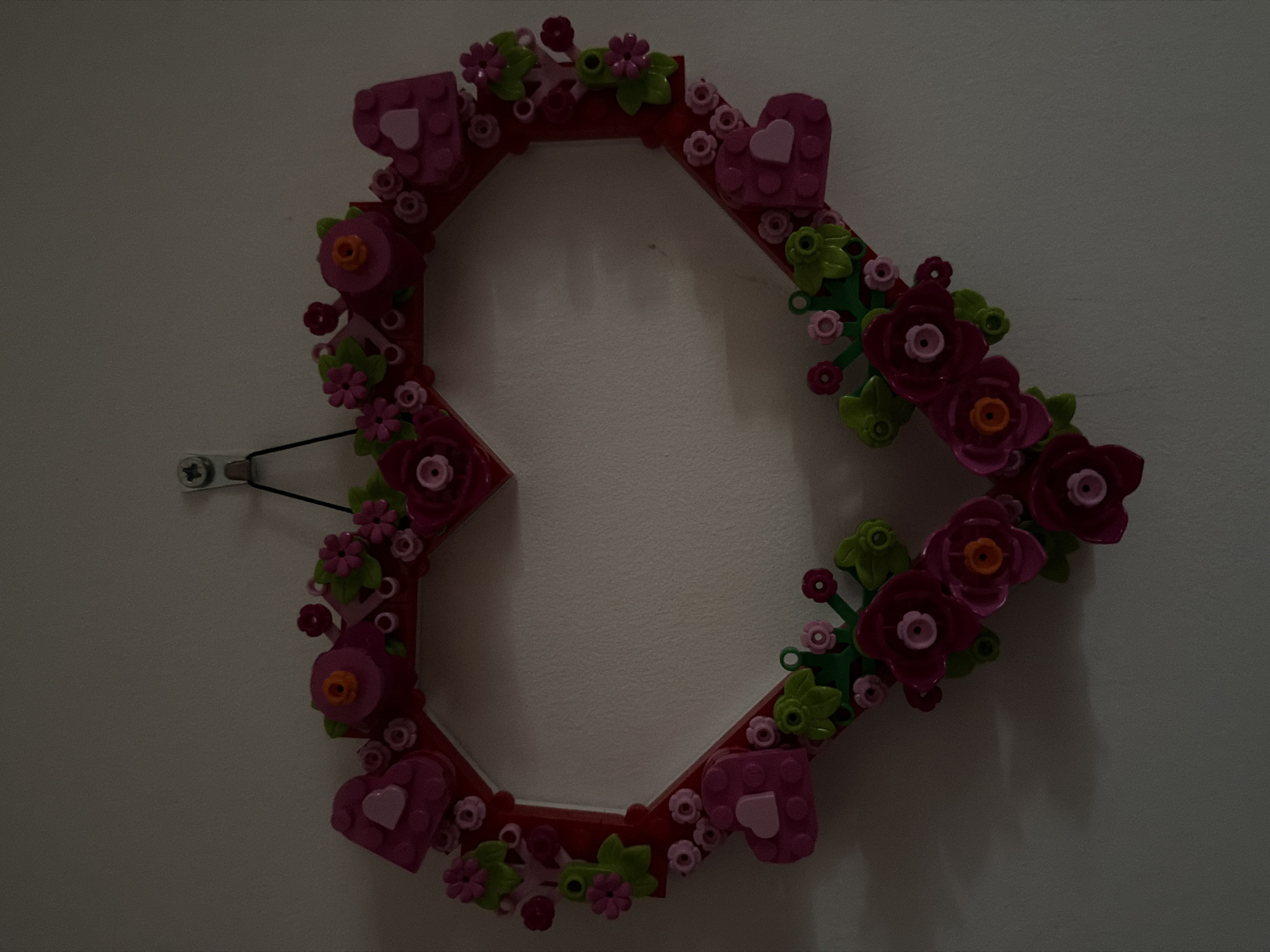 A heart-shaped wreath, constructed from red and pink building blocks, is displayed on a lightly textured, off-white wall in an indoor setting. The wreath is adorned with various pink and red flowers, green leaves, and pink heart shapes, all made from similar block elements. The wreath is suspended by a black string attached to a silver hook on the wall. The lighting in the image is dim, casting shadows on the wall and the wreath, suggesting it might be nighttime or a dimly lit room. The specific location in Souni-Zanatzia, Cyprus, cannot be determined from the visual information provided.