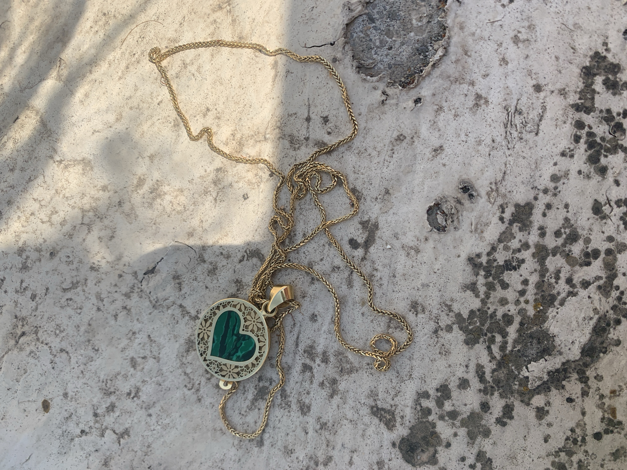 A gold chain necklace with a heart-shaped pendant rests on a textured, weathered concrete surface. The pendant features a green, malachite inlay shaped like a heart within a circular, ornate frame. The surface is mottled with darker patches and appears to be outdoors, with shadows cast from an unseen light source. The scene does not depict any specific location in Khazar Raion, Azerbaijan, nor does it include any people or actions.