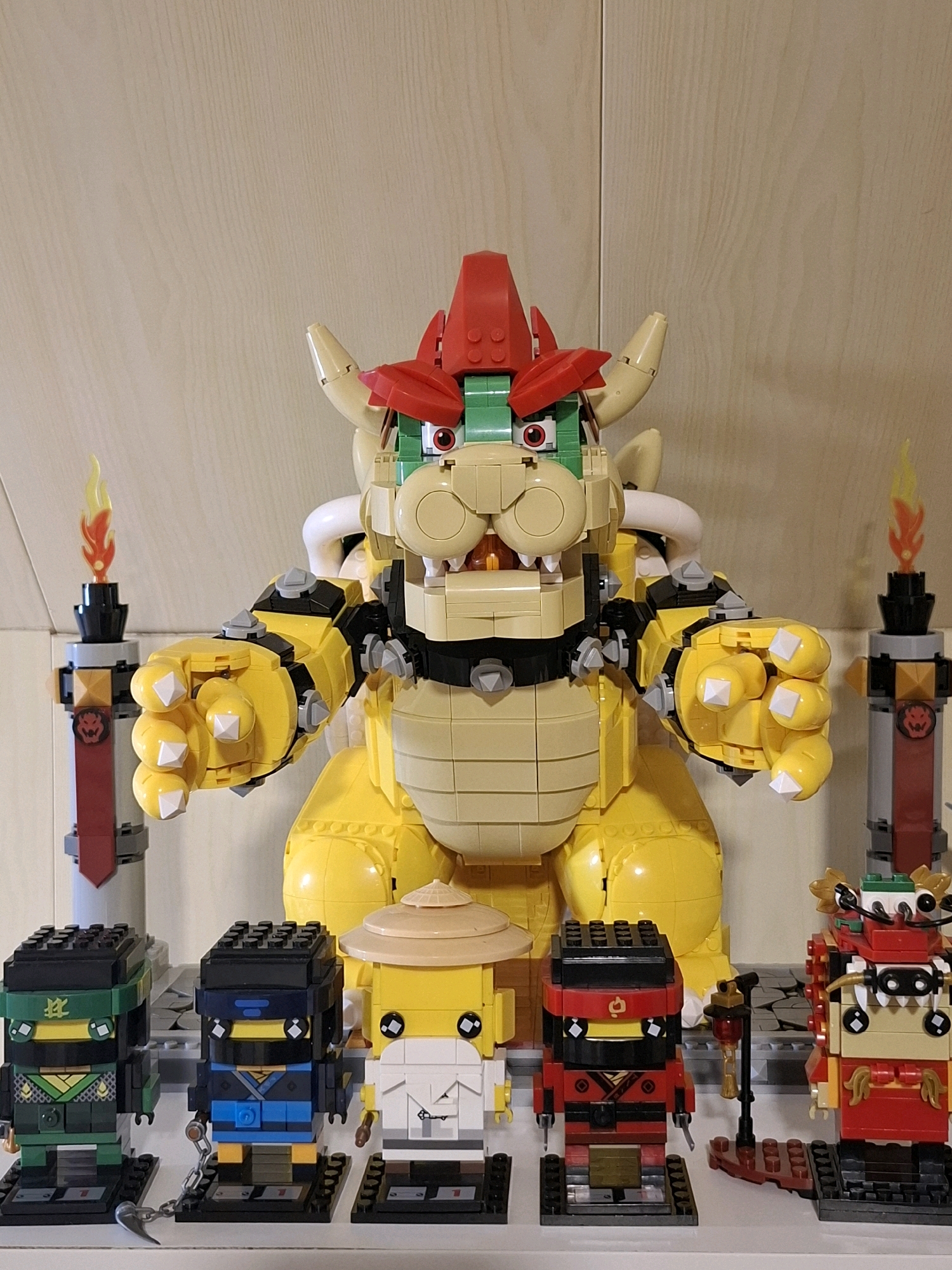 The image depicts a collection of Lego figures displayed on a shelf against a wood-paneled wall. In the center, a large, yellow Lego Bowser figure dominates the scene. To the left and right of Bowser are tall, gray Lego structures resembling torches with red banners and flame-like elements at the top. In front of Bowser, on the shelf, are several smaller Lego figures. From left to right, there are three figures resembling ninjas in green, blue, and red attire, followed by a yellow figure wearing a conical hat. To the far right, another Lego figure is visible, appearing to be a dragon or a similarly ornate character. The lighting suggests an indoor environment. There is no visible indication of the city of Tyumen, Russia, or any specific location within it. The scene is static and composed of assembled Lego models.