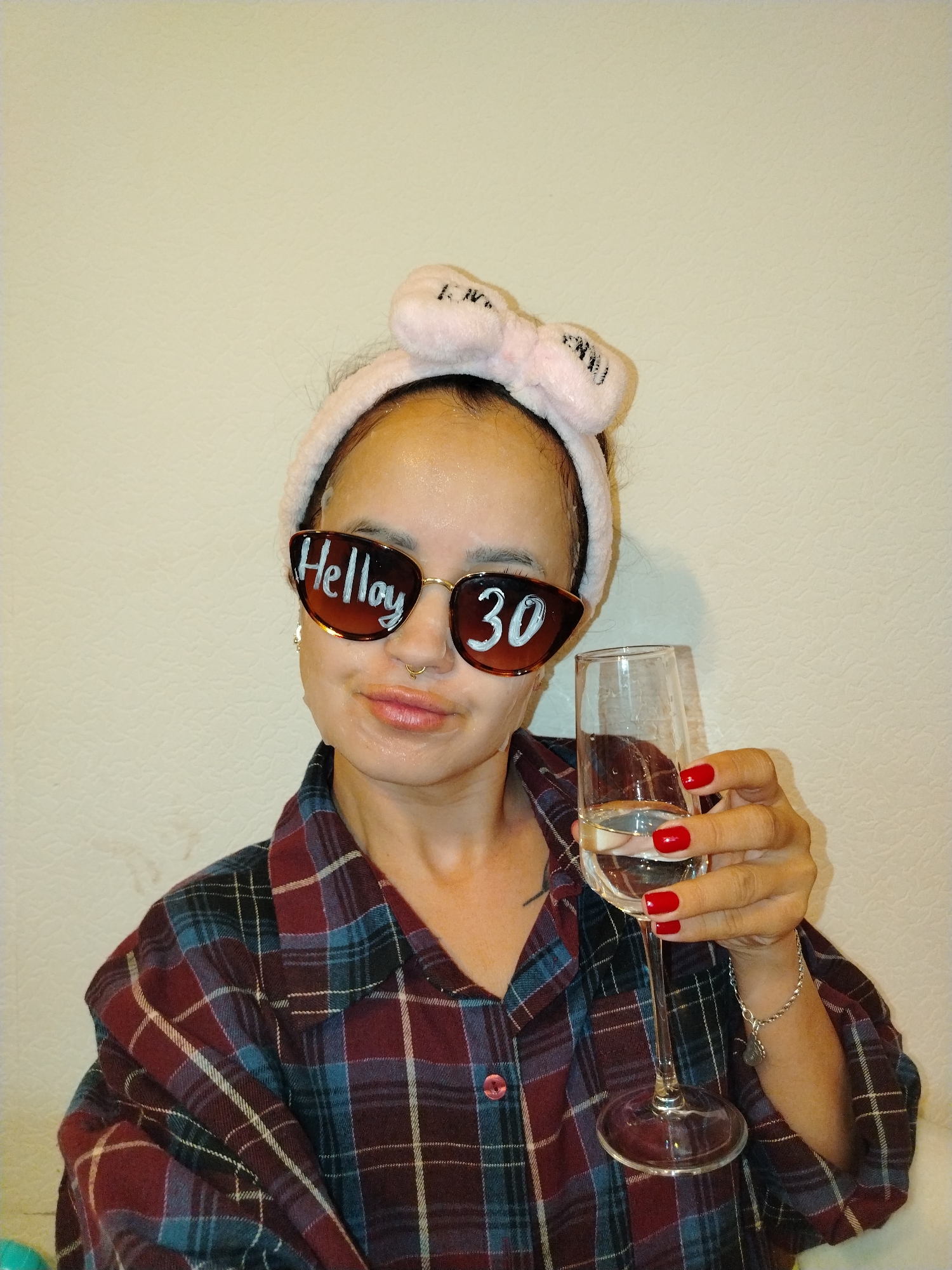 In the city of Noviy, Russia, a woman is depicted in a close-up shot, front-facing, holding a glass of clear liquid. She is wearing a pink, bow-adorned headband and large sunglasses with "Helloy 30" written on them in white. Her face is covered in a white sheet mask, and she has a nose ring. She is dressed in a dark plaid pajama top. Her fingernails are painted red, and she is wearing a silver bracelet on her right wrist. The background is a plain, textured off-white wall.