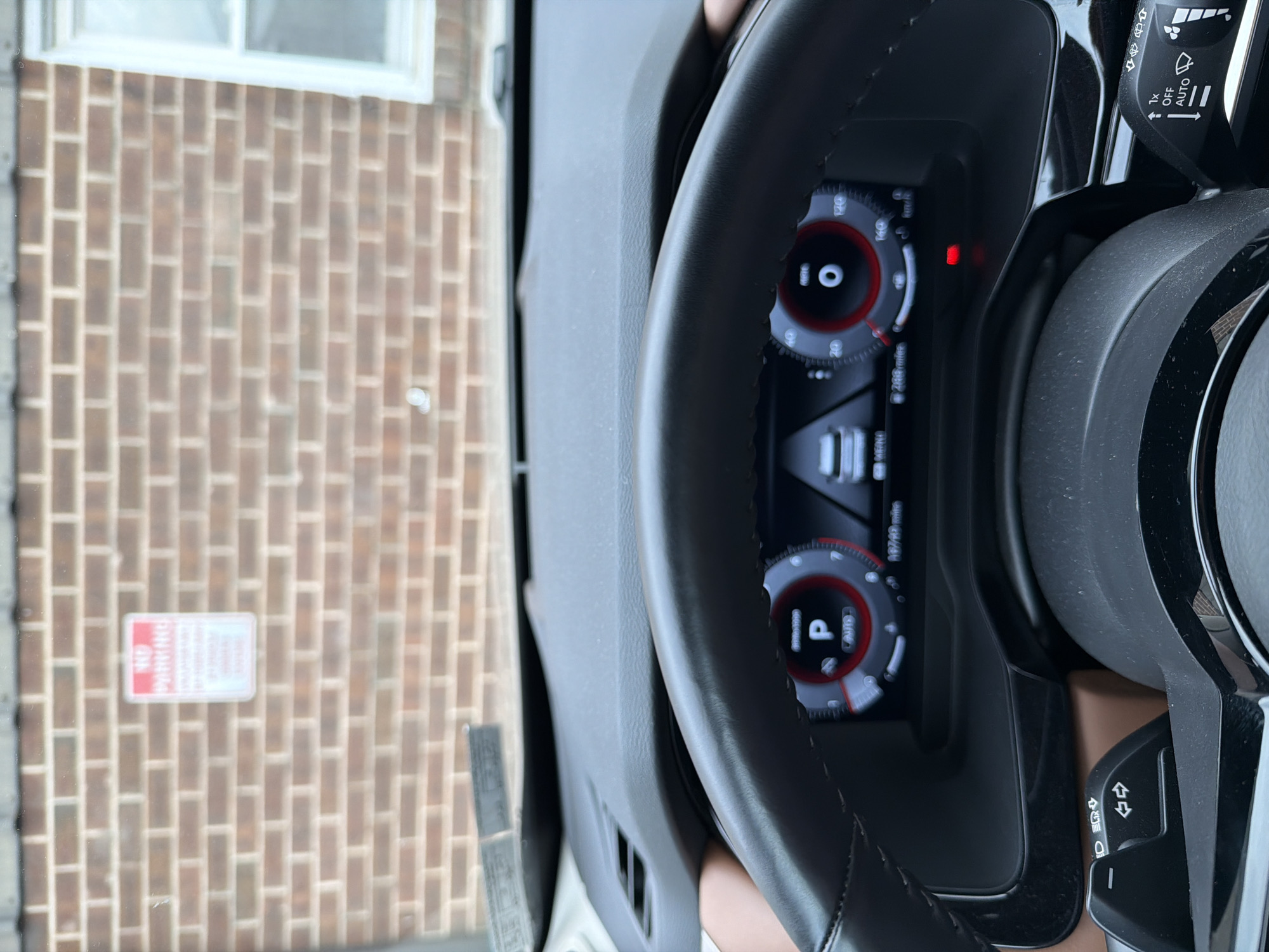 The image shows a close-up view of a car's interior, focusing on the dashboard and steering wheel. The dashboard features a digital display with various gauges and indicators. One gauge shows "10" with a red circle around it, suggesting a speed or RPM reading. Another display shows "P" indicating the car is in park. There's also a reading of "2400 miles." The steering wheel is black leather with controls on the right side, including a stalk with symbols for turn signals and headlights. The background is blurred, but appears to be an outdoor scene with a brick wall and a "NO PARKING" sign. The lighting suggests it is daytime. The overall scene is the interior of a parked car, with the exterior visible through the windshield. The image is oriented vertically, with the top of the car's interior at the bottom of the frame.