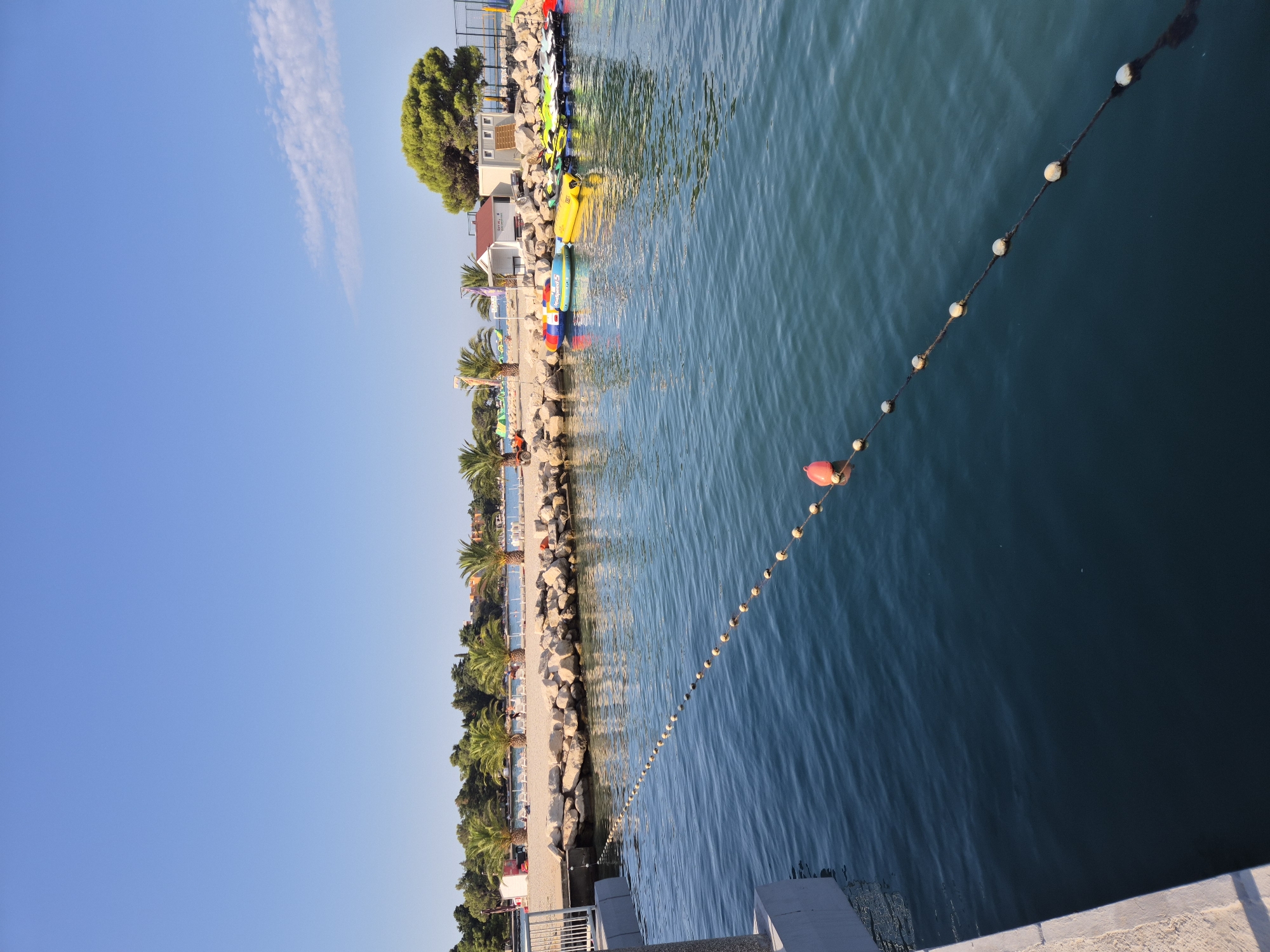 This image depicts a sunny day at a waterfront area in Umag, Croatia, likely a beach or marina. The foreground shows calm, dark blue water with a rope strung across it, marked by numerous small buoys and one larger orange buoy. To the left, a concrete walkway or pier edge is visible. The background features a rocky shoreline with a sandy beach beyond, lined with palm trees and some vegetation. Several beach chairs are set up on the sand. In the mid-ground, a collection of inflatable water toys and personal watercraft (jet skis) are parked near small, white buildings. A larger, mature tree stands prominently on the right side of the background, with a sports court visible behind it. The sky is clear and bright blue, indicating good weather. There are no people clearly identifiable in the image.