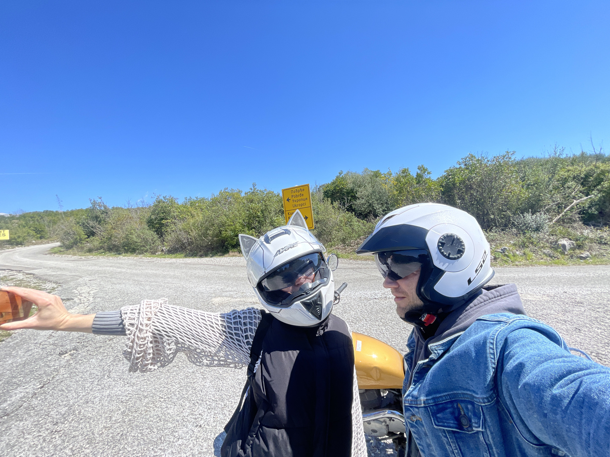 Two individuals, wearing motorcycle helmets, are positioned next to a motorcycle on a roadside in Kubasi, Montenegro. The person on the left wears a white helmet with cat ears and a mesh sweater and is holding a smartphone extended outwards as if taking a selfie or video. A road sign with Cyrillic writing is visible in the background. The sky is clear and blue. The environment appears to be a mountainous region with sparse vegetation.