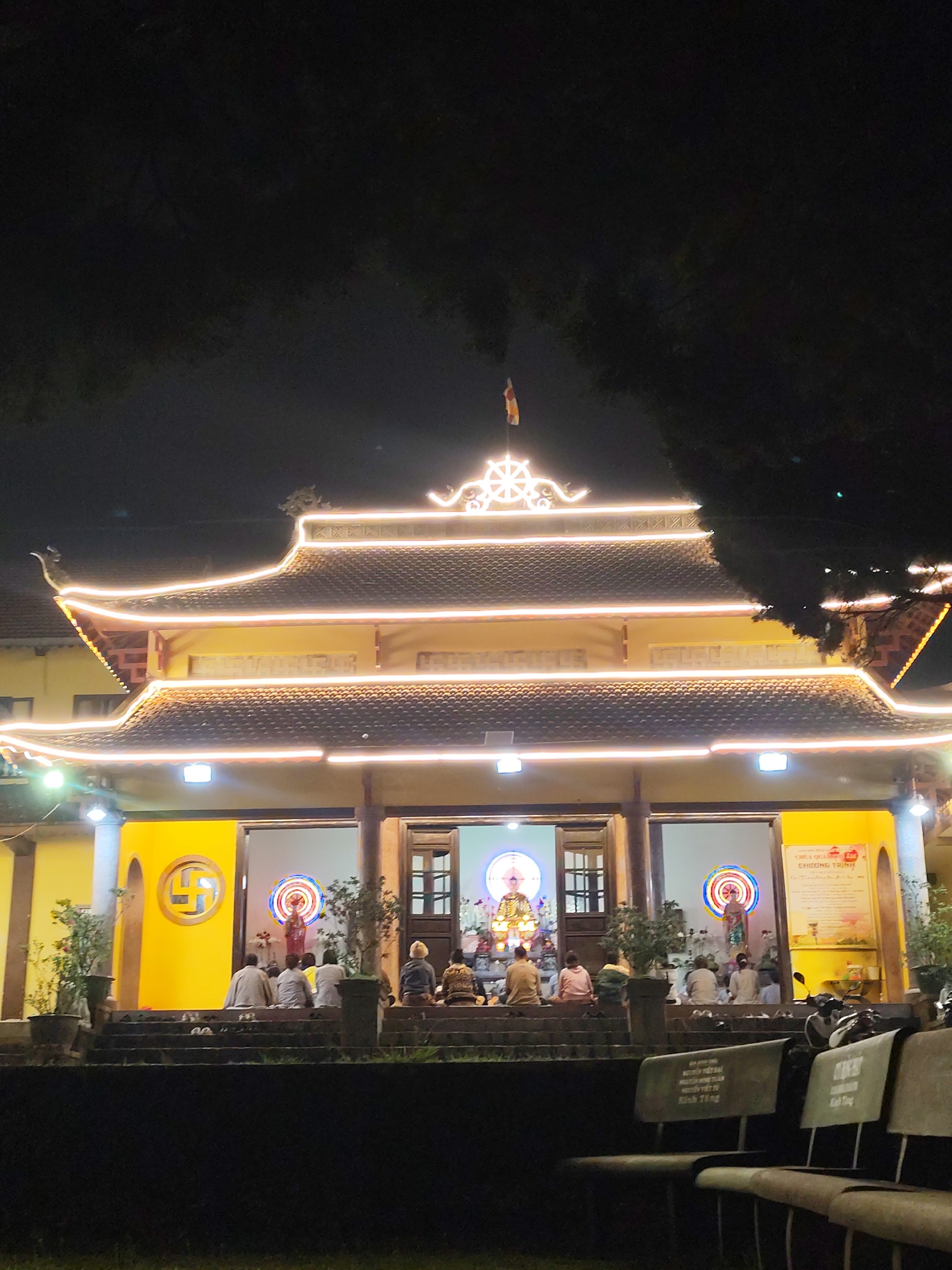 The image depicts a traditional Buddhist temple or pagoda at night in Da Lat, Vietnam. The multi-tiered structure is extensively decorated with bright string lights that outline its rooflines and architectural features, giving it a warm, inviting glow against the dark night sky. A prominent Dharma Wheel symbol adorns the peak of the roof, topped by a small flag, likely a Buddhist flag.
The temple's facade is a vibrant golden-yellow, with an open entrance revealing an illuminated interior. Inside, approximately 10-15 individuals are seated on the steps and floor, facing inwards towards brightly lit altars. At least two prominent statues, including what appears to be a golden Buddha and another deity, are visible within the temple, serving as focal points for the gathering. The attendees are dressed in simple clothing, indicating a religious ceremony or communal event. Shoes are neatly arranged on the steps in front of the seated individuals.
In the immediate foreground, several simple, light-colored chairs or benches are visible, also facing the temple. Some Vietnamese text is faintly legible on these chairs, such as "NGUYEN VIET DAI" and "Kinh Tụng," possibly dedications or instructions. On a sign inside the temple's right section, the Vietnamese words "CHƯƠNG TRÌNH" (Program) are clearly visible, suggesting a schedule for the event. Potted plants flank the temple entrance, adding to the serene ambiance. Dark foliage from trees frames the upper parts of the image.