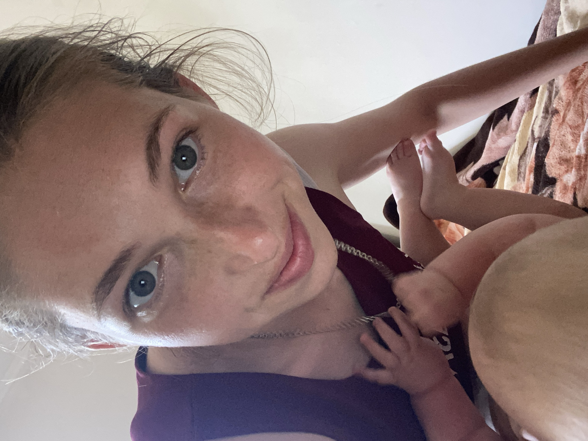 An extreme close-up, low-angle selfie shows a young woman with light skin and freckles looking directly at the camera. She has light brown hair, pulled back loosely. Her eyes are green, and she is wearing a dark red tank top, a silver chain necklace, and a light-colored bra underneath. The woman is holding a baby, whose head is in the foreground, and whose legs and feet are visible between the woman's chest and arms. The baby has light skin and appears to be wearing white socks or booties. The background is a plain, light-colored wall and a blanket with a brown and tan abstract pattern. The lighting is bright, suggesting daylight, and the overall impression is that the photo was taken indoors.