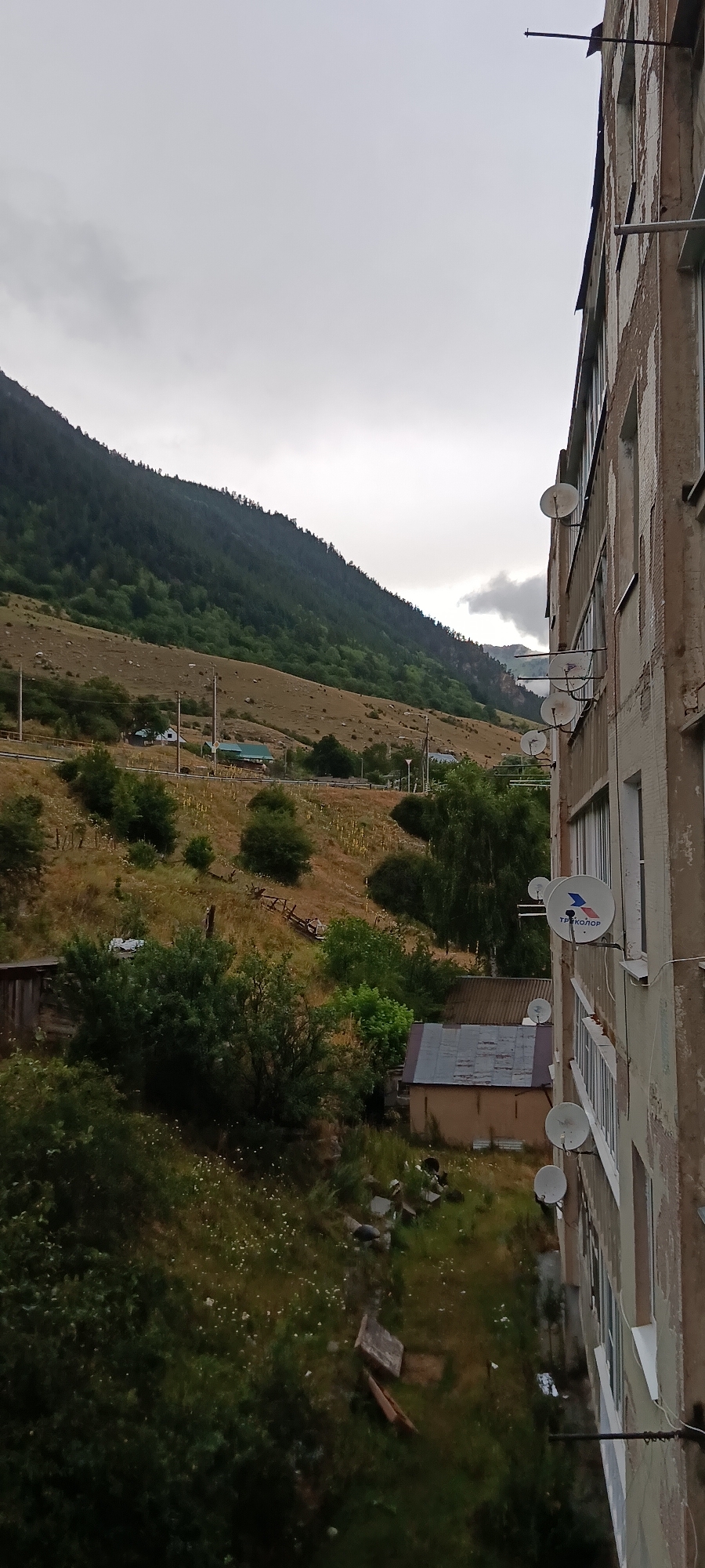 An outdoor, overcast, eye-level shot shows an apartment building on the right side of the frame and a hillside with trees and some buildings in the background. The apartment building is a pale beige structure with visible signs of wear and tear, and several satellite dishes are mounted on its exterior wall. The hillside is covered with dry grass and sparse vegetation, with a dense forest on the upper slope. A few small buildings, one with a green roof, are visible scattered on the hillside. The sky is cloudy and a muted grey. The scene appears to be a residential area in the town of Teberda, Russia, nestled in a mountainous region.