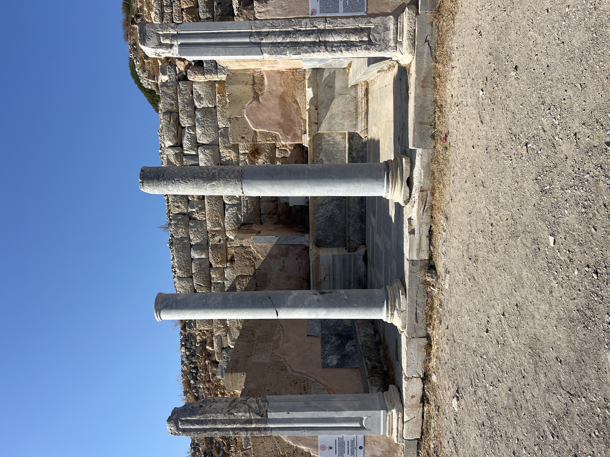 In an outdoor archaeological setting, four ancient marble columns stand prominently in the foreground, aligned horizontally. Two large, fluted columns with simple bases and capitals are centrally positioned, flanked by two narrower, more ornate columns on either side. These outer columns exhibit decorative carvings and stepped bases. Behind the columns, a rough stone wall forms the backdrop, constructed from irregular blocks of varying sizes and colors. The wall appears to be part of an ancient structure, with some sections showing signs of decay and reconstruction. The ground in front of the columns is unpaved, consisting of loose gravel and dirt. The sky above is a clear, vibrant blue. The scene is illuminated by bright sunlight, casting shadows on the ground and accentuating the textures of the ancient stonework.