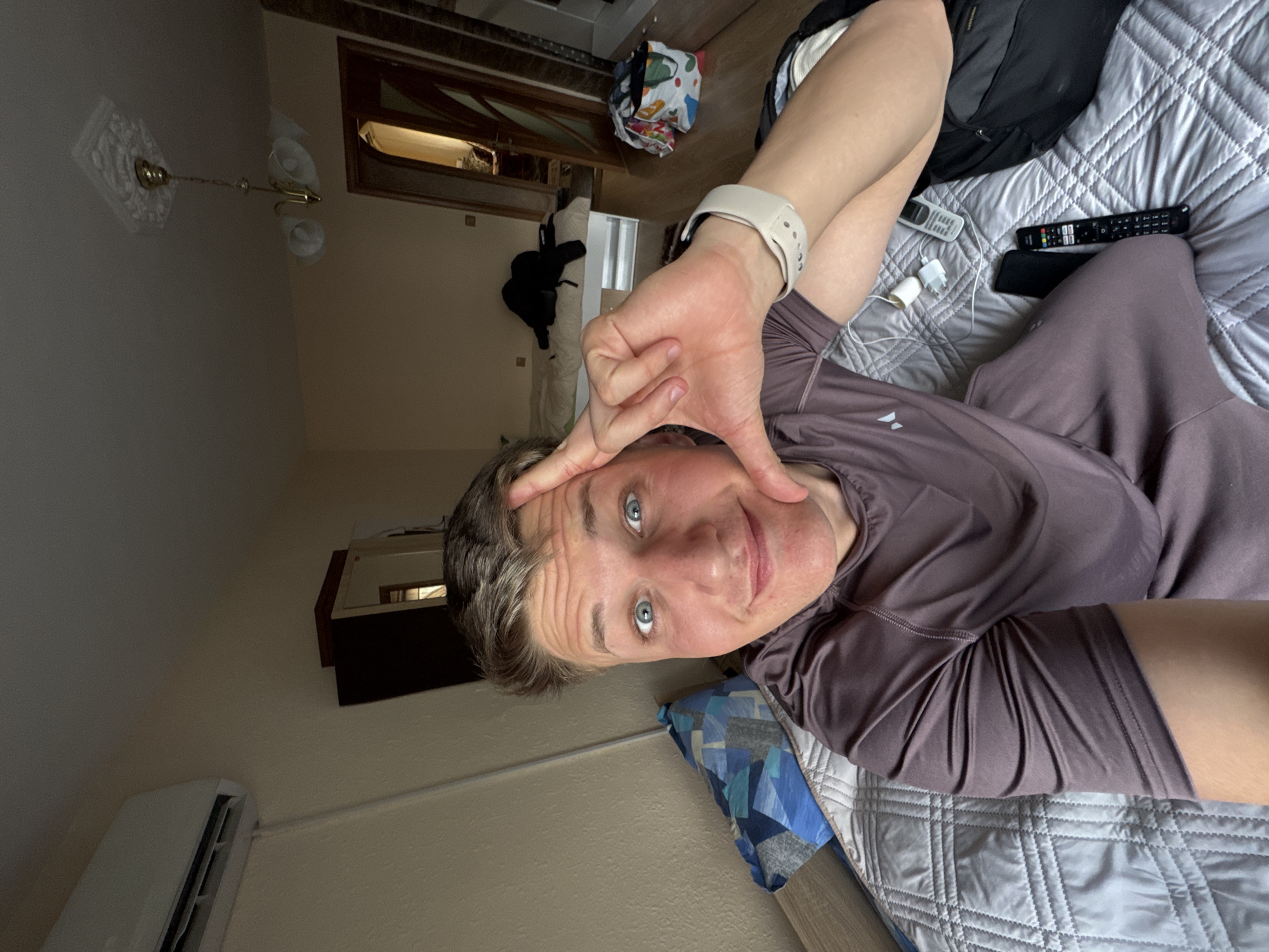 A selfie of a man with blue eyes is taken in a room. The man has short, light brown hair and is wearing a brown athletic shirt and a white smartwatch on his left wrist. He is lying on a silver quilted bed with a blue patterned pillow behind him. His left hand is raised, framing his face. A black backpack, a black smartphone, a white smartphone charger, and two TV remotes are on the bed beside him. In the background, there's a door leading to a darker room with a white chair. A ceiling light fixture is visible. An air conditioning unit is mounted on the wall. The location is likely inside an apartment or hotel room in Burgas, Bulgaria, though specific confirmation requires additional visual clues or context.