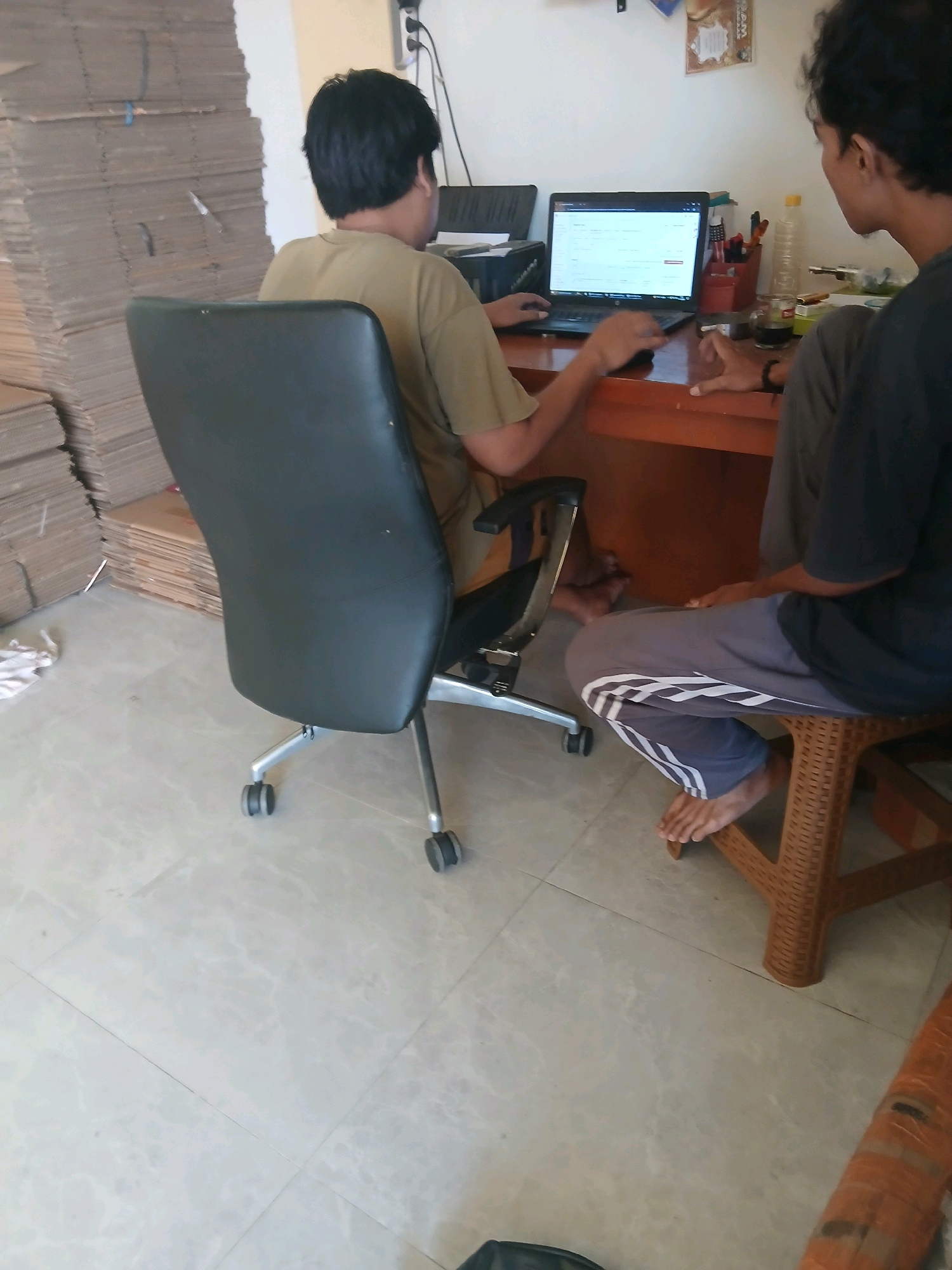 In Jepangpakis, Indonesia, two individuals are seen in what appears to be a storage or office area. One person is seated in a black office chair, facing a laptop on a wooden desk. Their hands are on the keyboard, suggesting they are actively using the computer. Next to the desk, a stack of large cardboard boxes is piled high against a wall. The second individual is seated on a brown woven stool, facing the desk from the right side of the frame. They are wearing athletic pants with white stripes. On the desk, a laptop is open, displaying what looks like a spreadsheet or web page, along with a cup of dark liquid, a bottle of water, and a red container filled with pens and other small items. The floor is tiled with a light-colored, patterned material. The environment seems to be indoors, with a plain white wall in the background.