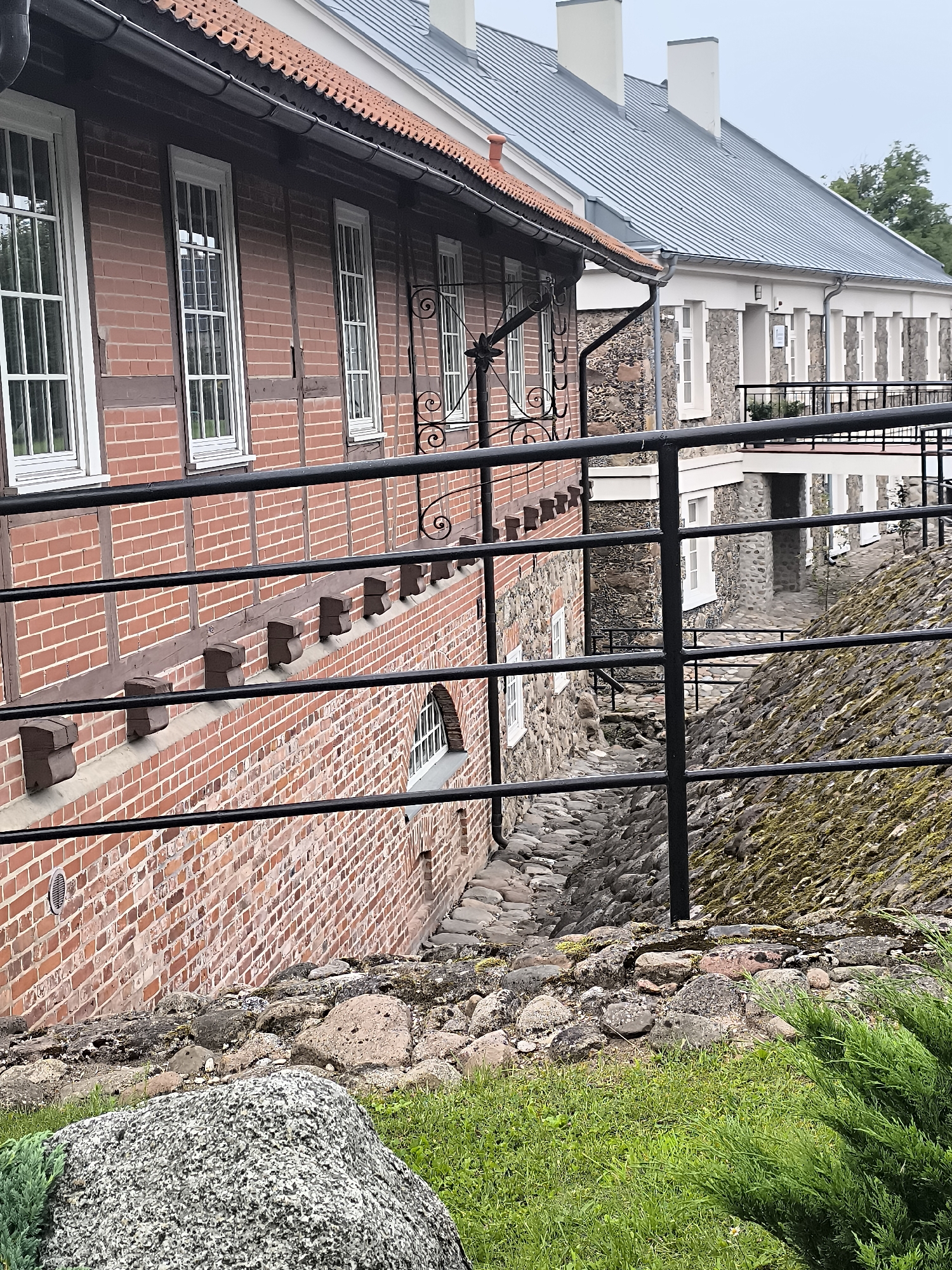 The image captures a section of the Kretinga Manor complex in Kretinga, Lithuania. The foreground is dominated by a large, grey, speckled boulder, lush green grass, and a dark green, bushy plant. A black metal railing runs horizontally across the midground, obscuring the view of the manor's lower levels. Beyond the railing, a weathered brick wall with dark timber framing forms the side of a building. This wall features several tall, white-framed windows. Adjacent to the brick structure, a stone-faced building with a grey, corrugated metal roof is visible. This building also has white-framed windows and a balcony with a white railing. The ground between the buildings is a cobbled pathway descending downwards, bordered by a moss-covered stone embankment on the right.