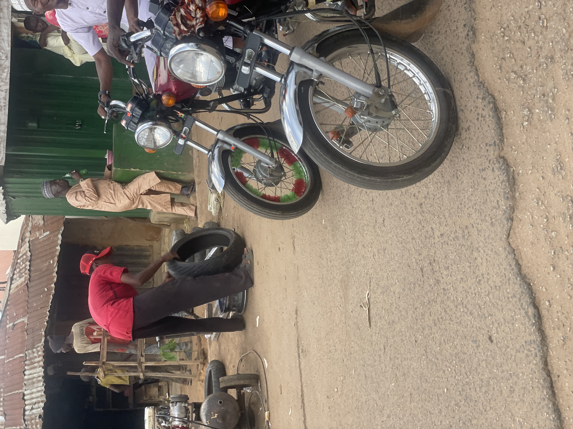 In Jos, Nigeria, a man in a red shirt and baseball cap is working on a motorcycle tire. He's holding the tire and appears to be removing it. Another man in a light-colored traditional outfit is sitting nearby, holding a green bottle. Two motorcycles are parked close together, one with a red fuel tank and the other with a maroon fuel tank. The scene is outdoors, with a corrugated metal structure and a dusty ground visible. There's a sense of activity and a busy, perhaps informal, workshop setting. The time of day appears to be daylight, with no strong indicators of specific weather conditions.