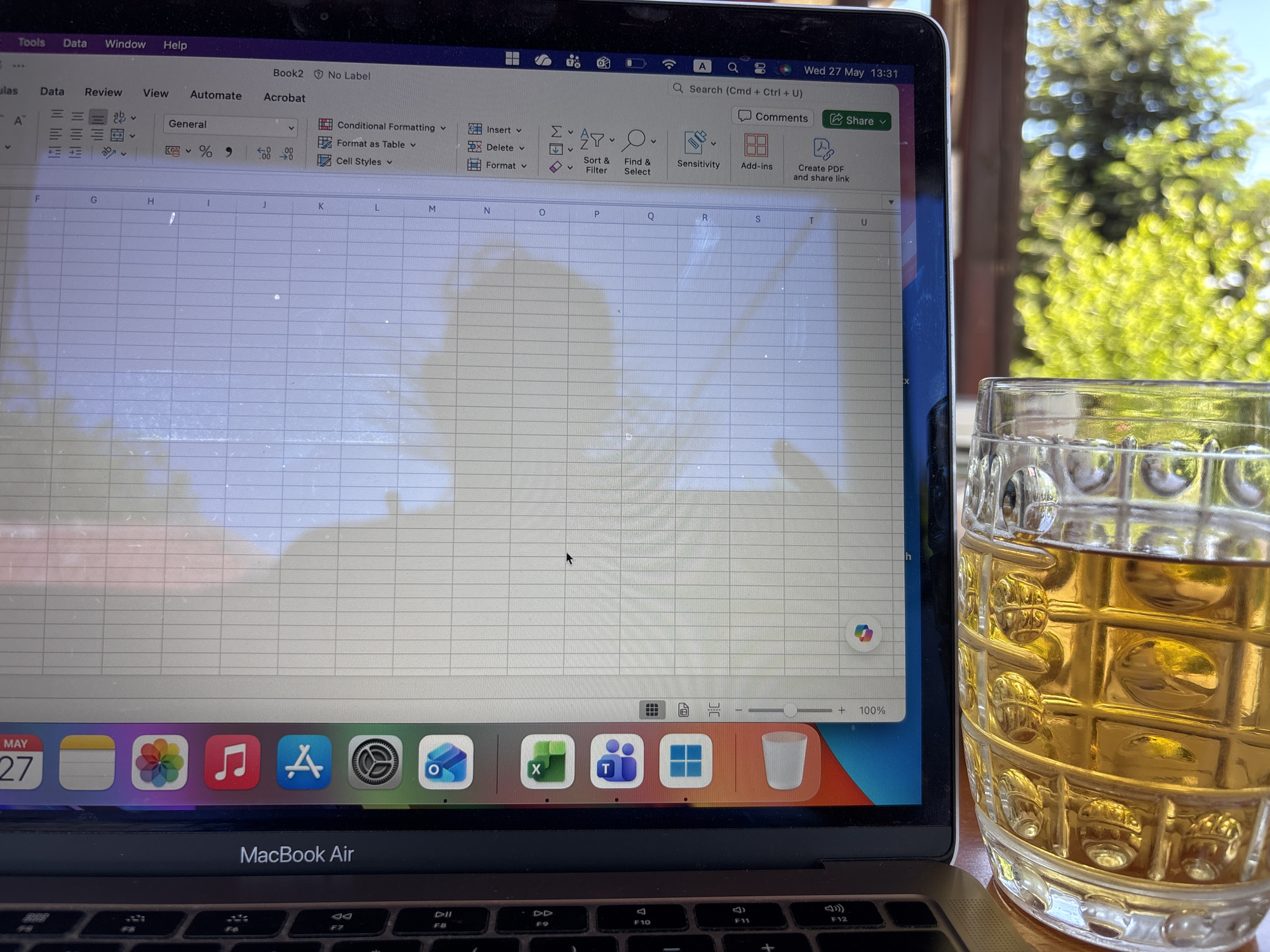 The image shows a MacBook Air open, displaying a spreadsheet program, likely Excel, with "Book2 - No Label" at the top. The date "Wed 27 May 13:31" is visible in the upper right corner of the screen. Below the screen are application icons in the dock, including Calendar, Photos, Music, Safari, Settings, Outlook, Excel, Teams, and Windows. A glass mug filled with a golden liquid, possibly beer or iced tea, sits to the right of the laptop. The background is blurred, showing green foliage and hints of sunlight, suggesting an outdoor setting. The image likely depicts a work-from-home situation with a beverage nearby. There is no information to suggest that the picture was taken in the city of Pchelnik, Bulgaria.