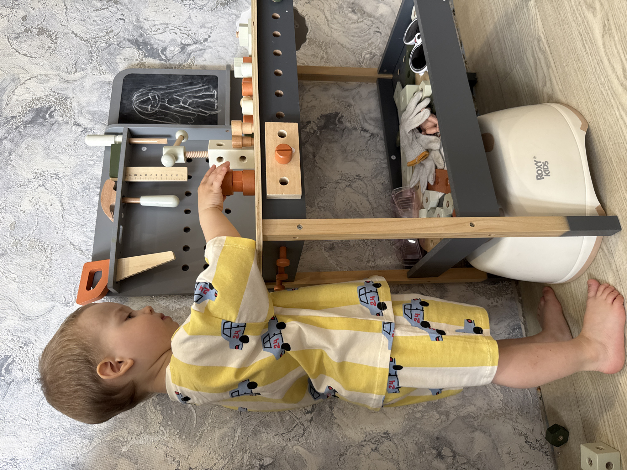 A toddler in a yellow and white striped shirt with blue car patterns lies on a gray and white patterned floor. The child's head is near the left side of the frame, and their feet extend toward the lower right. The child is interacting with a gray and wood-colored toy workbench which has various tools and accessories. A white baby potty with "Roxy Kids" printed on its side is positioned to the right of the workbench. A wooden block toy sits near the child's feet in the lower right corner of the image. The environment has a flat, even light. This photo was taken in the city of Rusanovo, Russia.