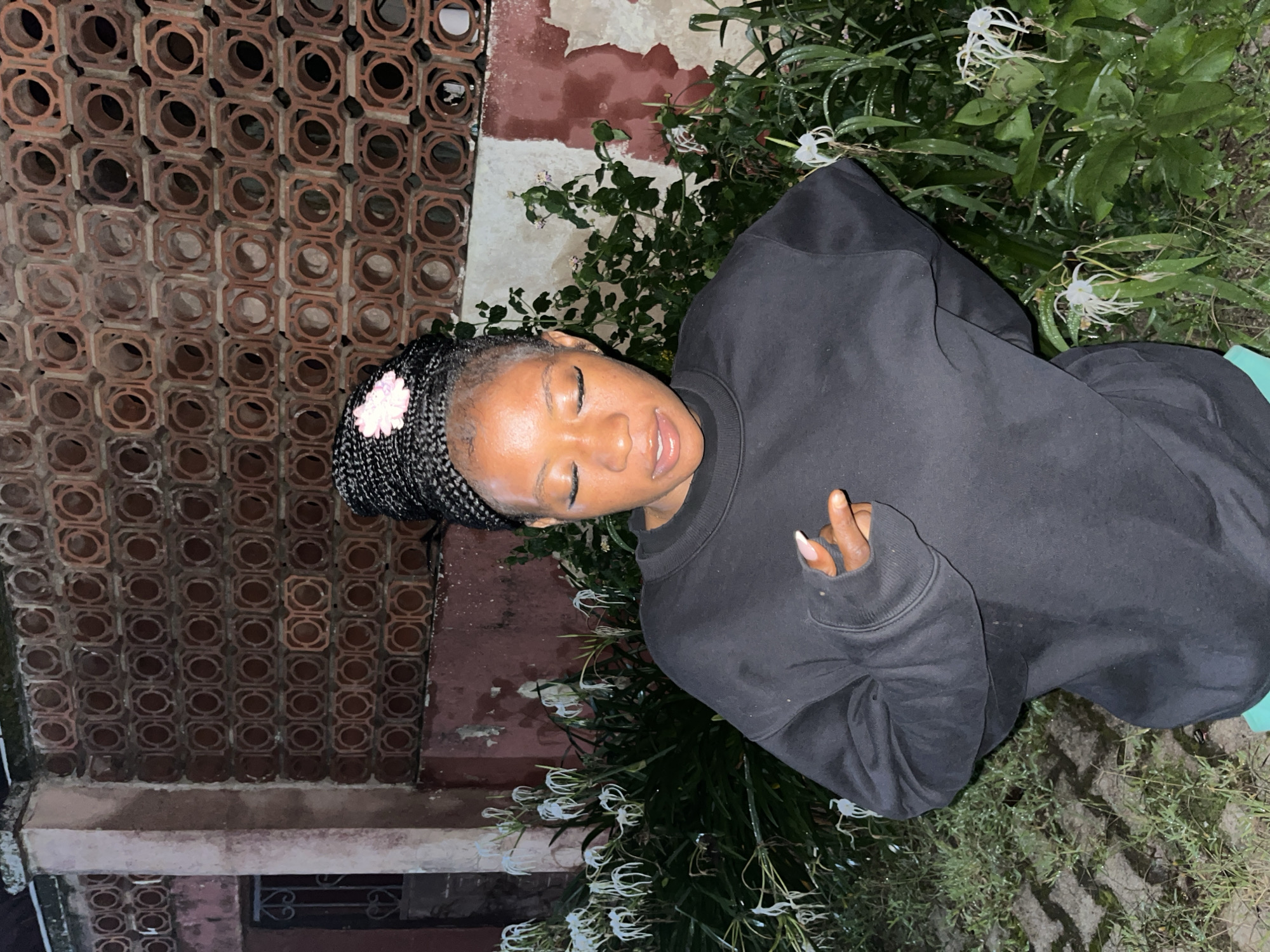 A young Black woman stands outdoors at night, posing with her eyes closed and head tilted down. She has braided hair styled in a bun with a small pink flower accessory. She is wearing a black crewneck sweatshirt and appears to have on turquoise shorts. The background consists of a patterned brick wall with a distressed, pinkish-red finish and dense foliage with white, spidery flowers. The ground is a mixture of paved stones and grass, appearing damp. The setting is consistent with outdoor residential areas found in cities like Jos, Nigeria, where such architectural styles and flora are common.