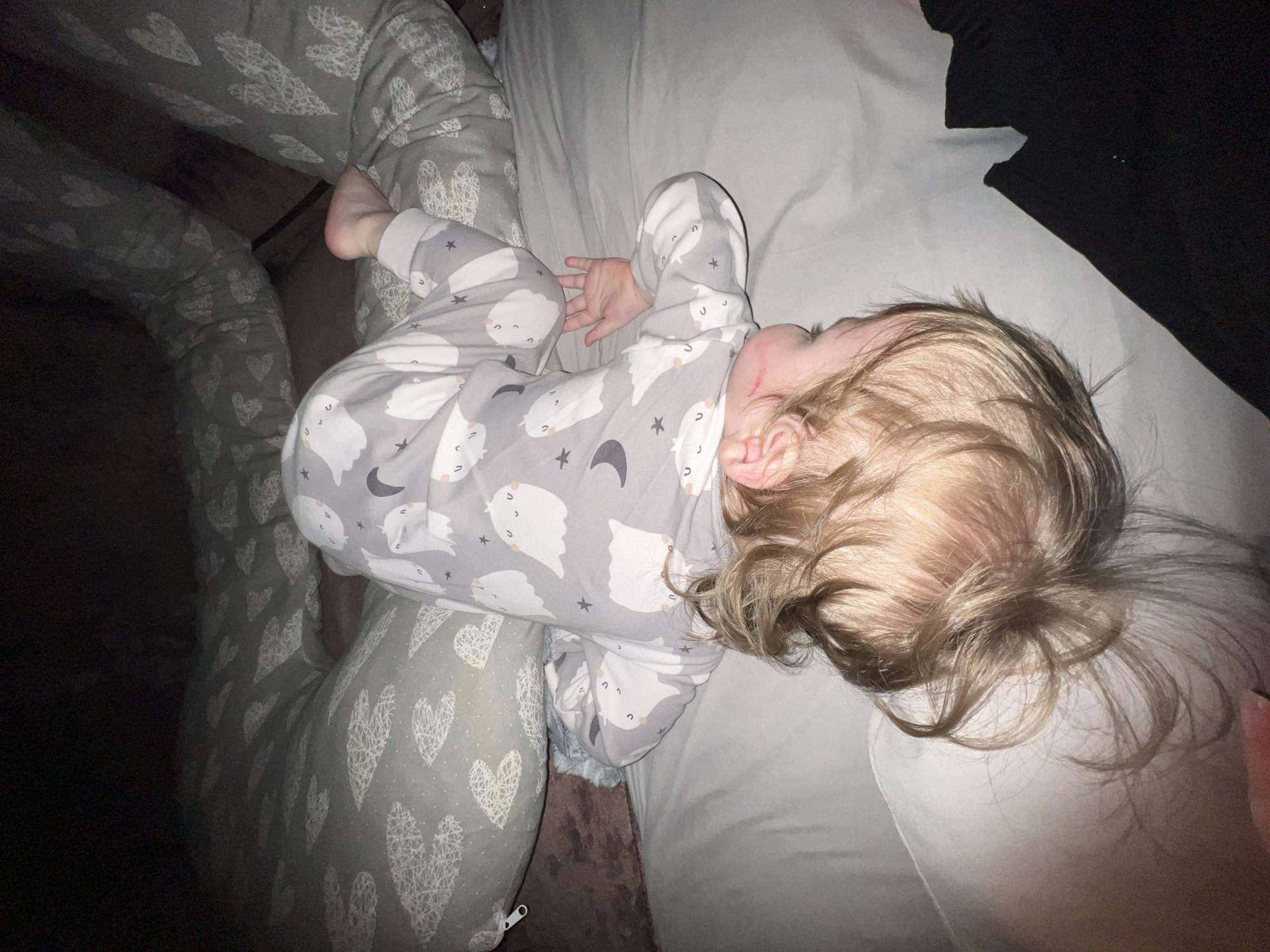 A young child lies asleep on a light gray bedsheet, wearing gray and white pajamas patterned with clouds, moons, and stars. The child is positioned on their side with their face turned away from the camera, revealing blond hair spread across the pillow. To the left of the child, someone is wearing gray pajamas with white heart patterns, presumably an adult. To the right, a portion of dark clothing is visible on the sheet. The location is likely indoors, indicated by the lighting and lack of outdoor elements. The mention of Noviy, Russia, is unsupported by the visual details of the image.
