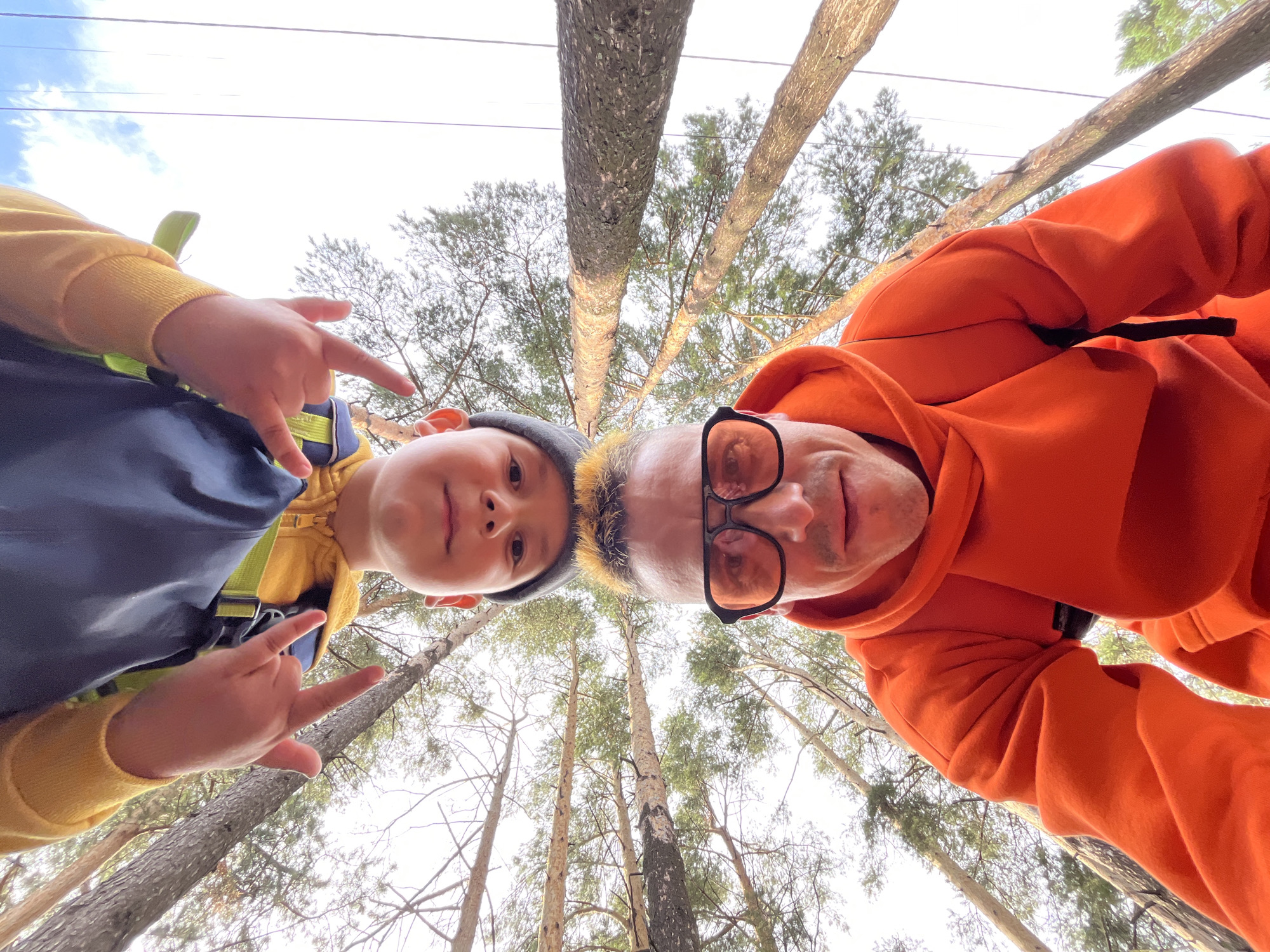 An outdoor, eye-level, wide-angle, low-angle selfie taken in Chaikovsky, Russia, features two people in the foreground with trees and a bright sky in the background. The subjects are oriented upside down. In the upper portion of the frame, a young person wearing a yellow hooded jacket over a dark blue garment and a grey beanie is visible. The person is making a hand gesture with their index and pinky fingers extended upwards. Below this person, a man with blonde-tipped hair, wearing an orange hooded sweatshirt and oversized orange-tinted glasses, is looking towards the camera. The background consists of tall, slender pine trees with sparse branches and needles, reaching towards a pale, cloudy sky with some dark lines that could be power lines.